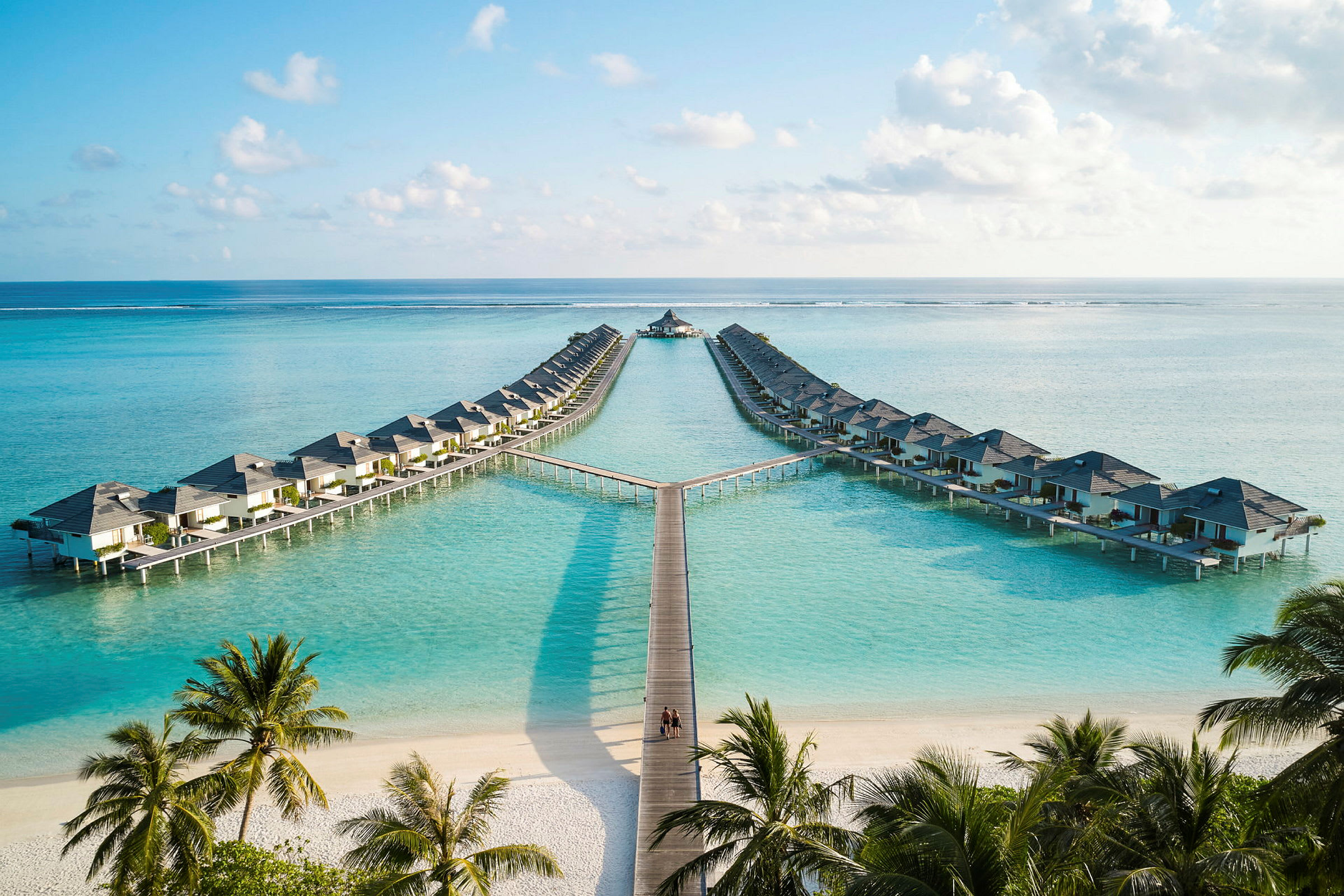 Hotellbungalower på påler over klart blått vann, med sandstrand og palmer i forgrunnen. (AI-generert tekst)