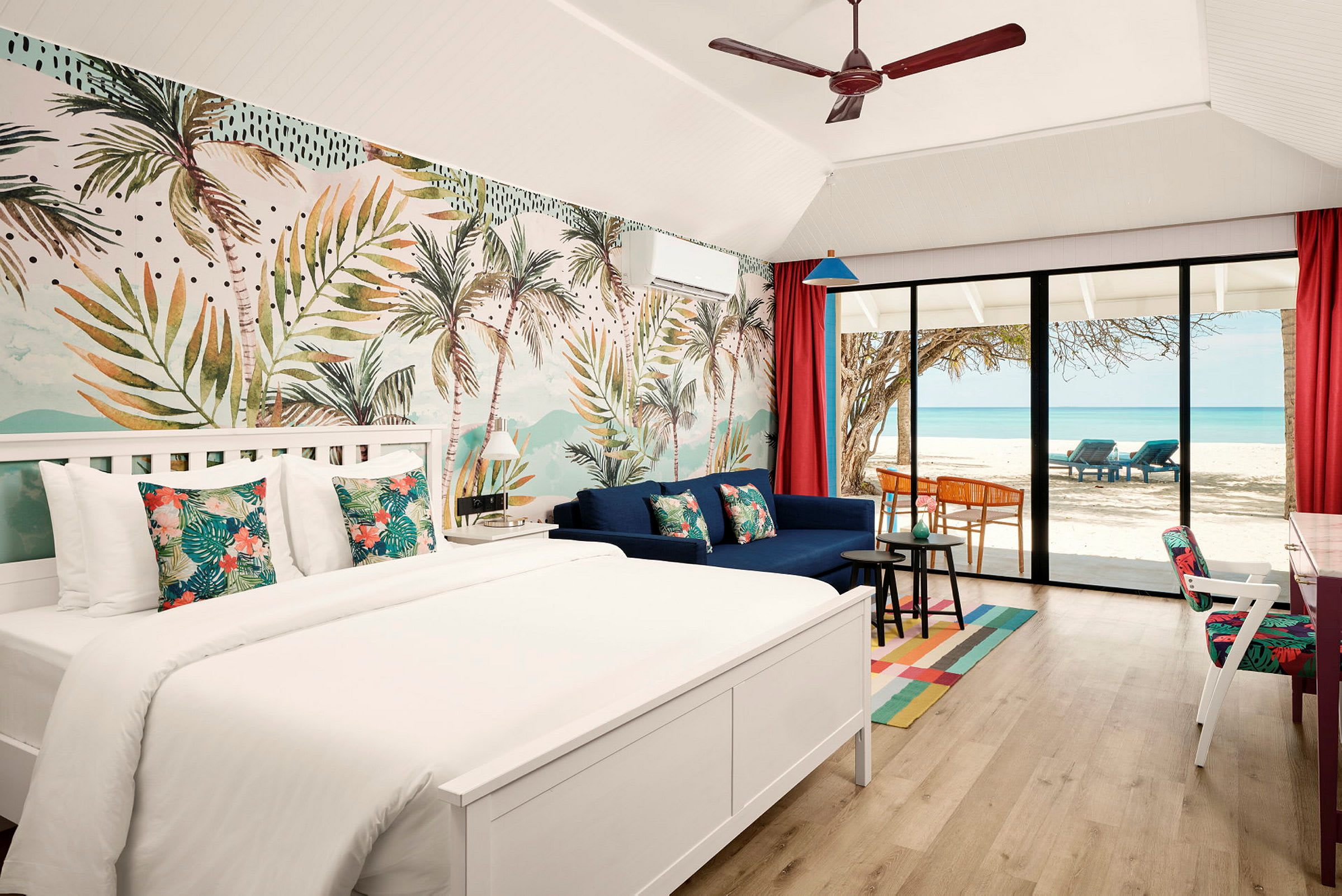Et hotellrom med en dobbeltseng, sofa og en stor glassdør som vender mot en strand med palmer og havutsikt. (AI-generert tekst)
