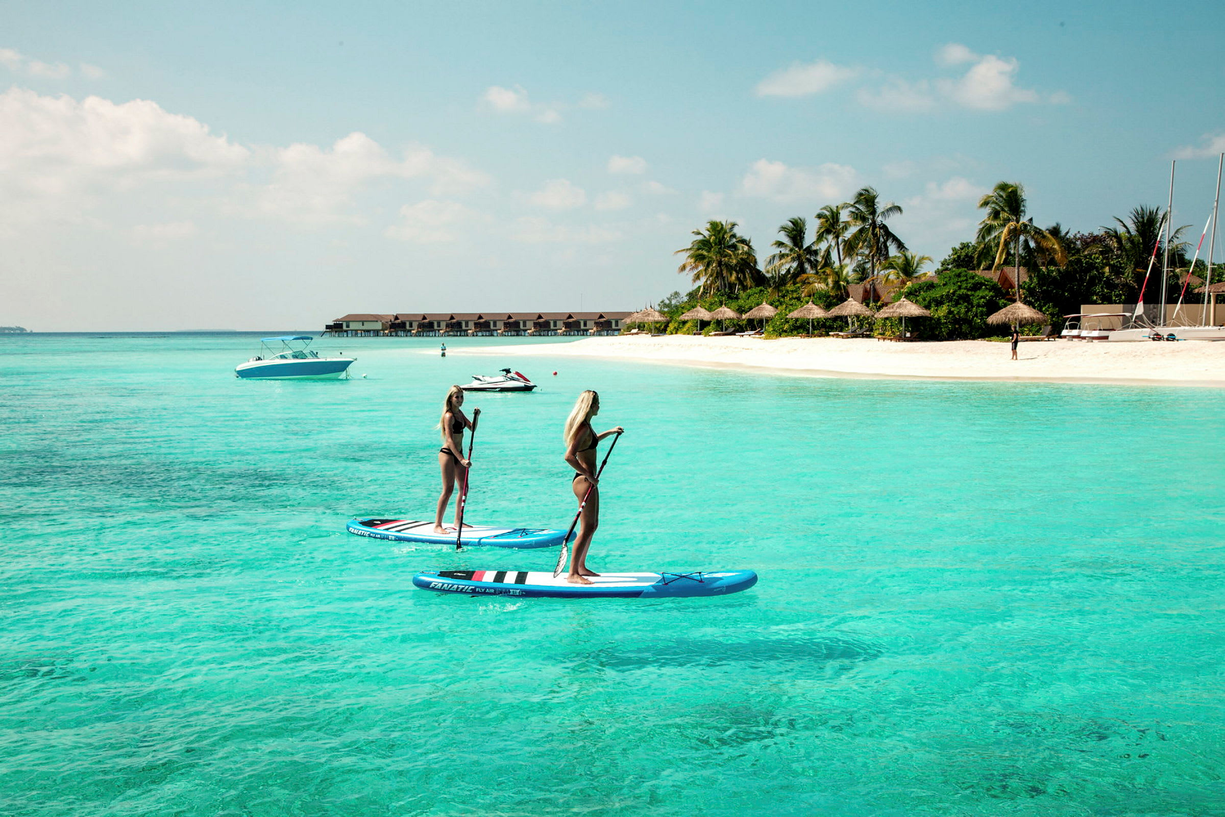 To personer står på paddleboards i klart turkist vann nær en sandstrand. (AI-generert tekst)