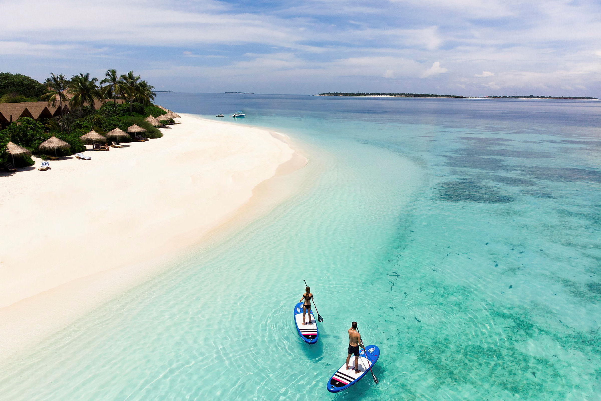 To personer står på paddleboards i klart turkist vann ved en sandstrand med hytter og palmer. (AI-generert tekst)