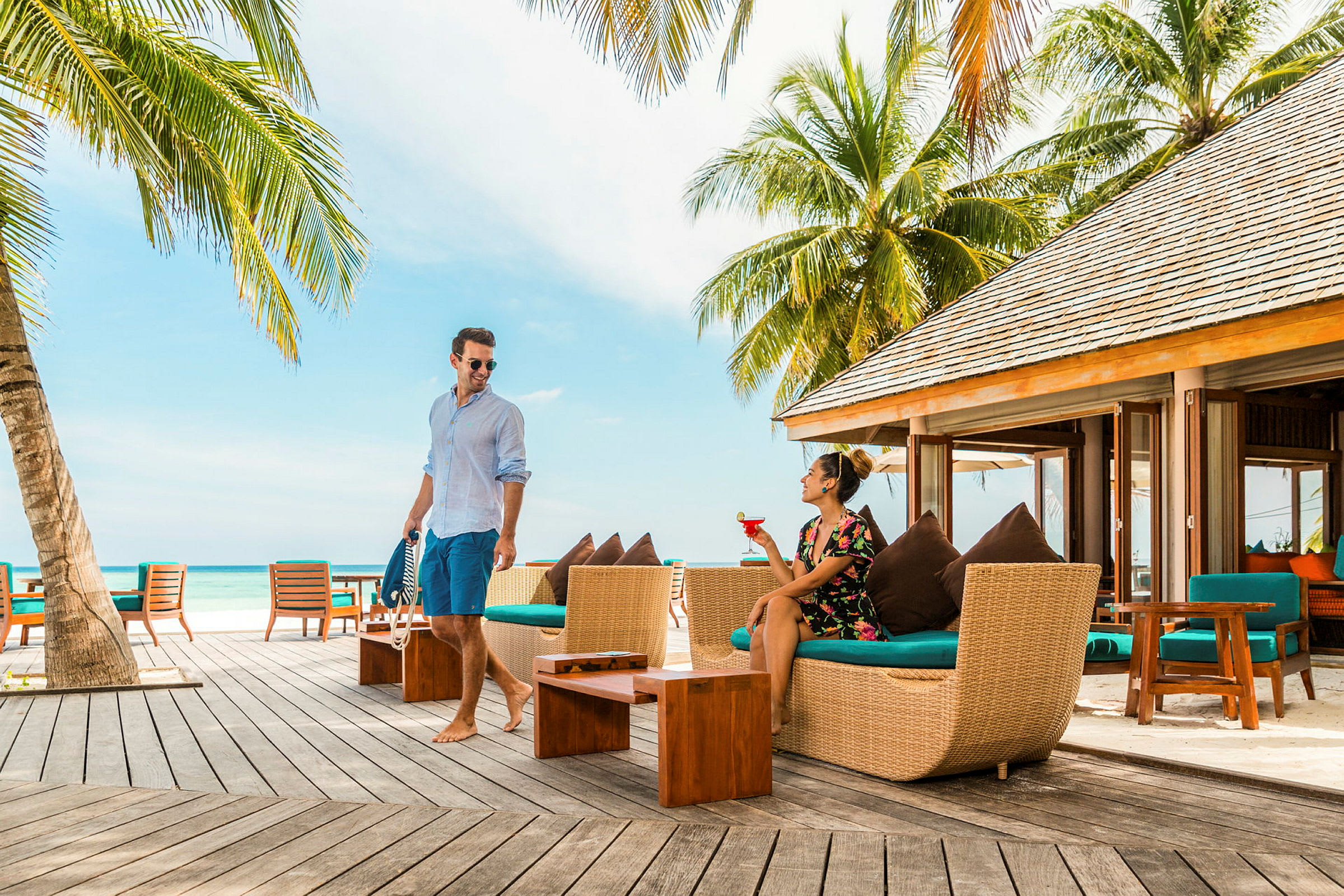 Et par personer nyter en utendørs lounge med palmer og havet i bakgrunnen. (AI-generert tekst)