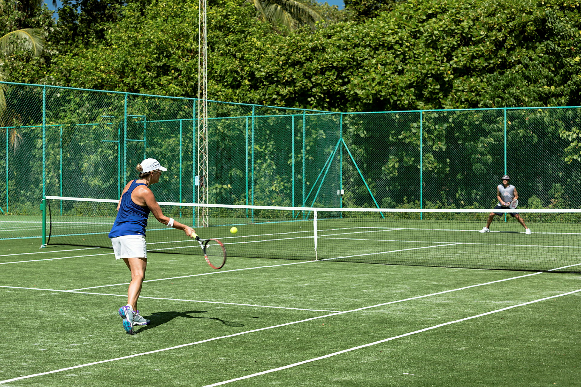En person spiller tennis på en grønn tennisbane omgitt av frodig natur. (AI-generert tekst)
