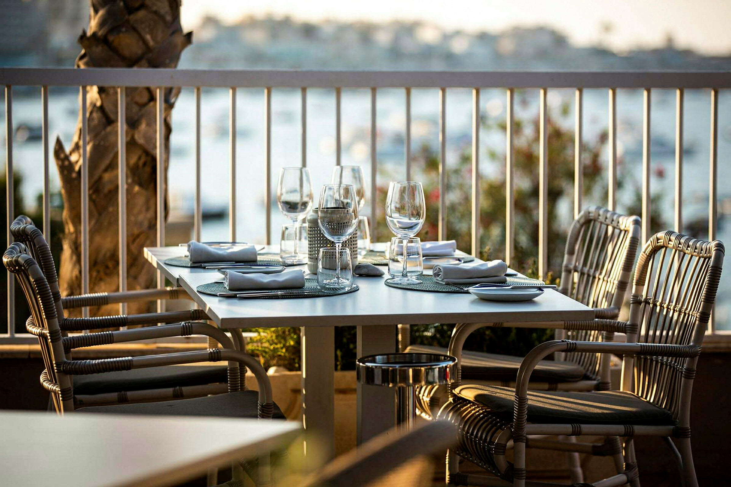 Et dekket bord med glass og servietter på en utendørs terrasse, omgitt av utsikt over vannet. (AI-generert tekst)