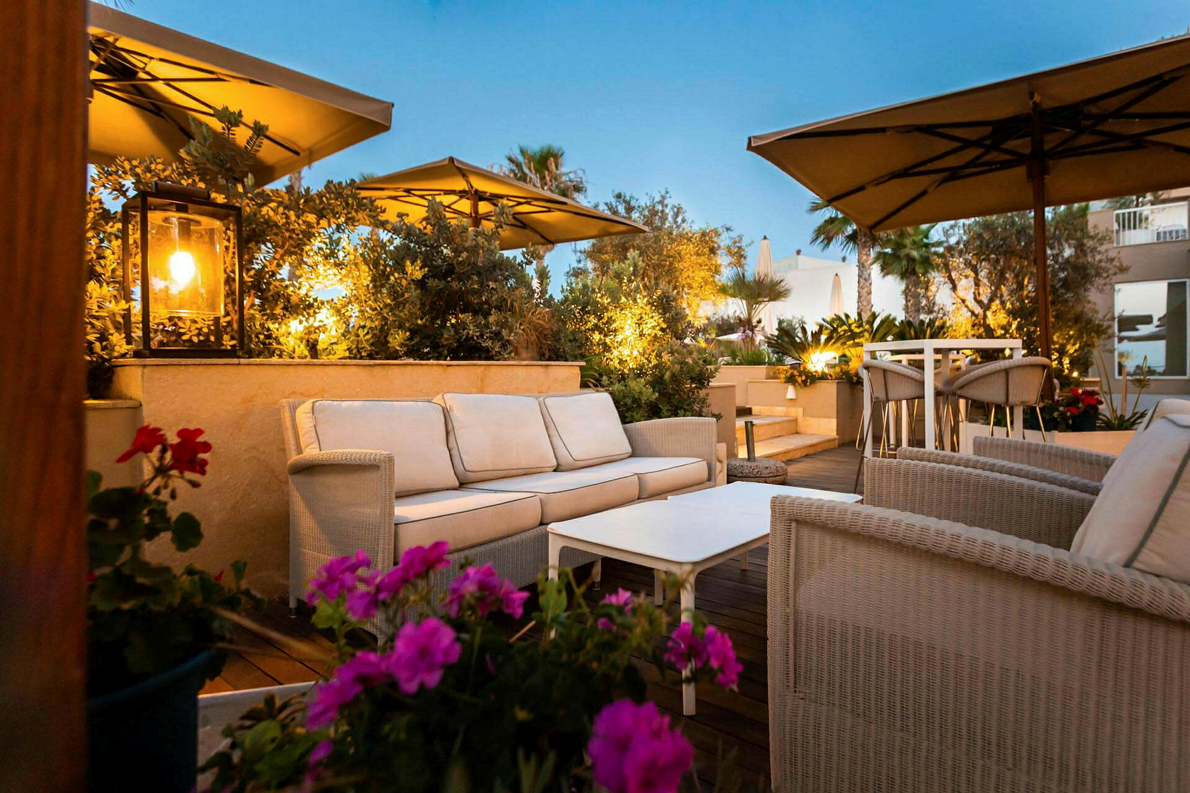 En terrasse med sittegrupper med kurvmøbler og parasoller, omgitt av planter og blomster. (AI-generert tekst)