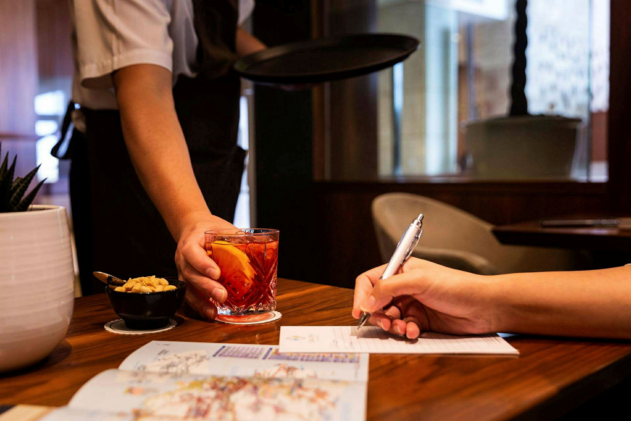 En servitør serverer en drink i et glass ved siden av en person som skriver på et papir. (AI-generert tekst)