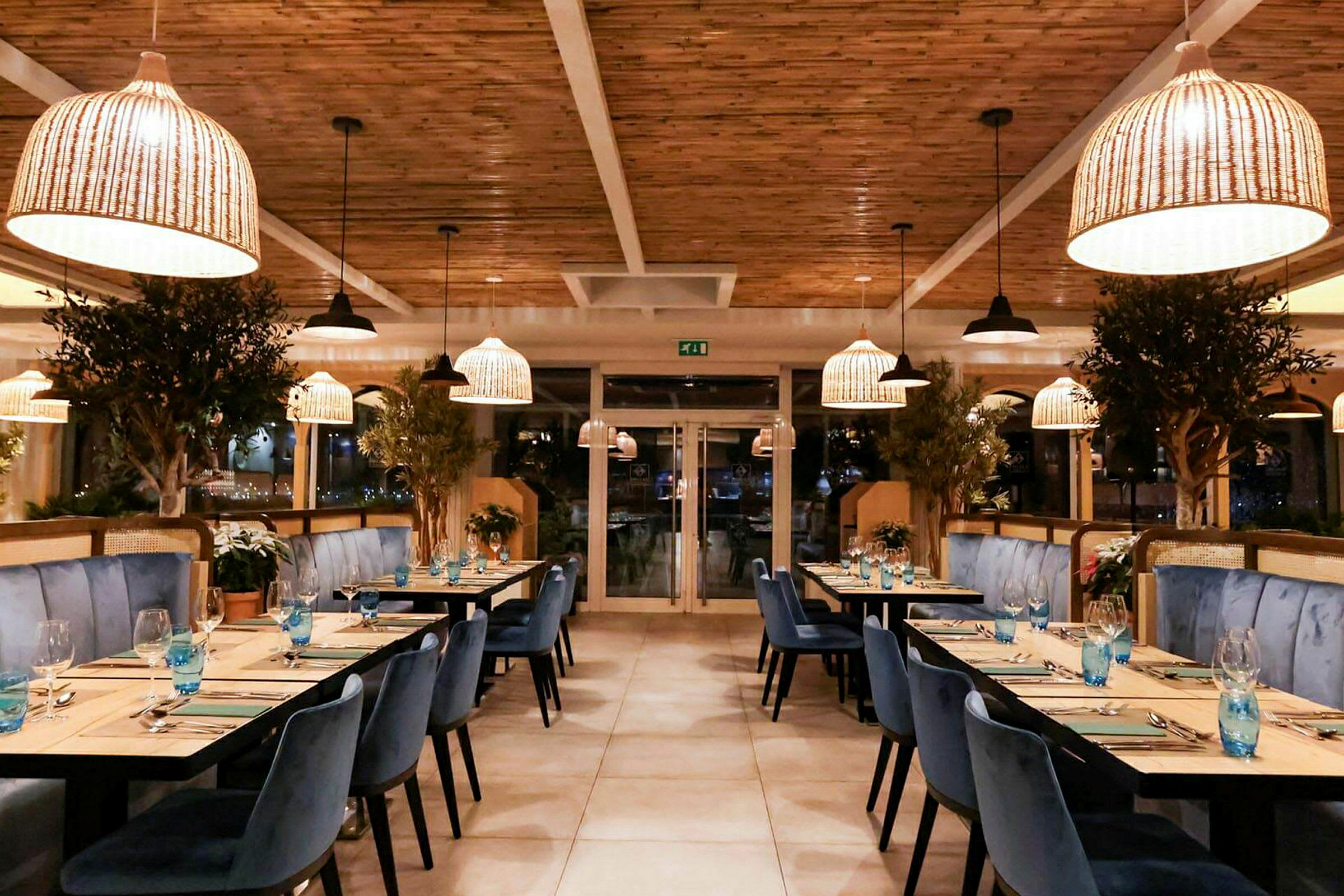 En elegant restaurantinteriør med blå stoler, trebord og store plantepynt. Rottinglamper henger fra taket. (AI-generert tekst)