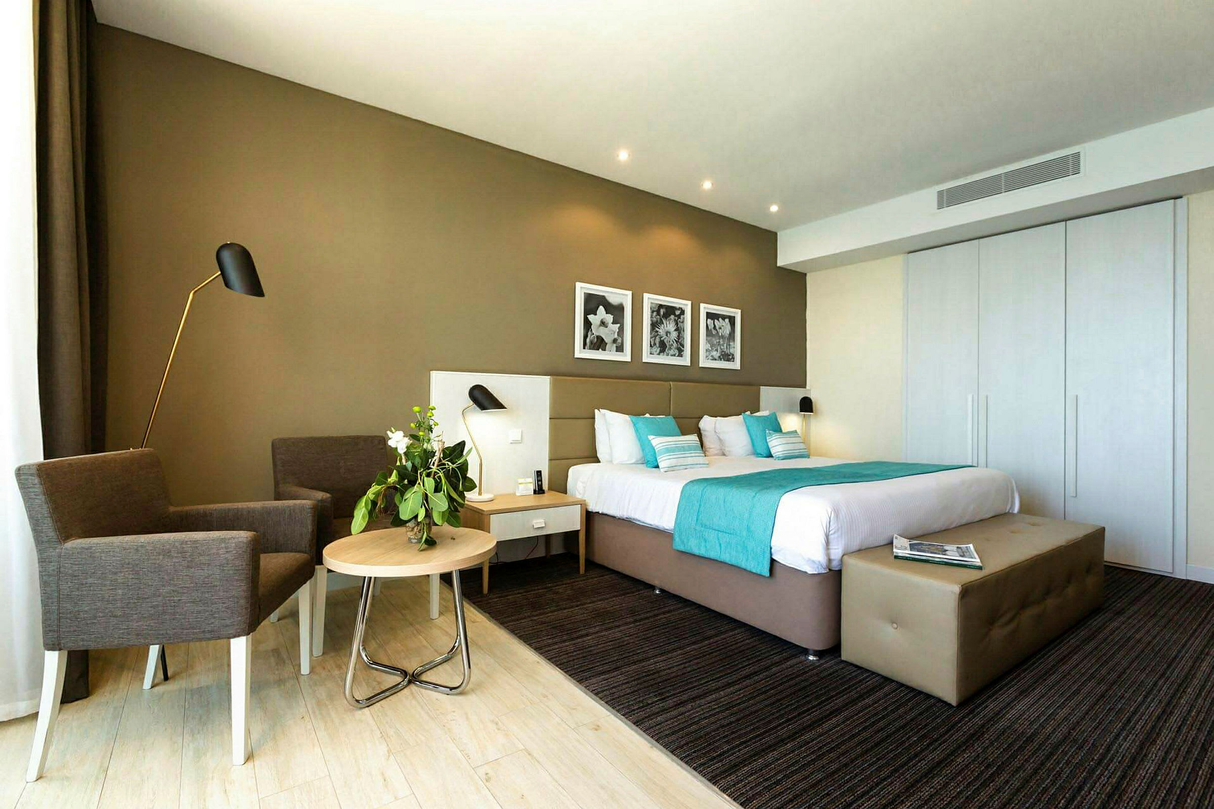 Et moderne innredet hotellrom med en stor seng, dekorative puter og et nattbord. Et par lenestoler står ved siden av et lite bord. (AI-generert tekst)