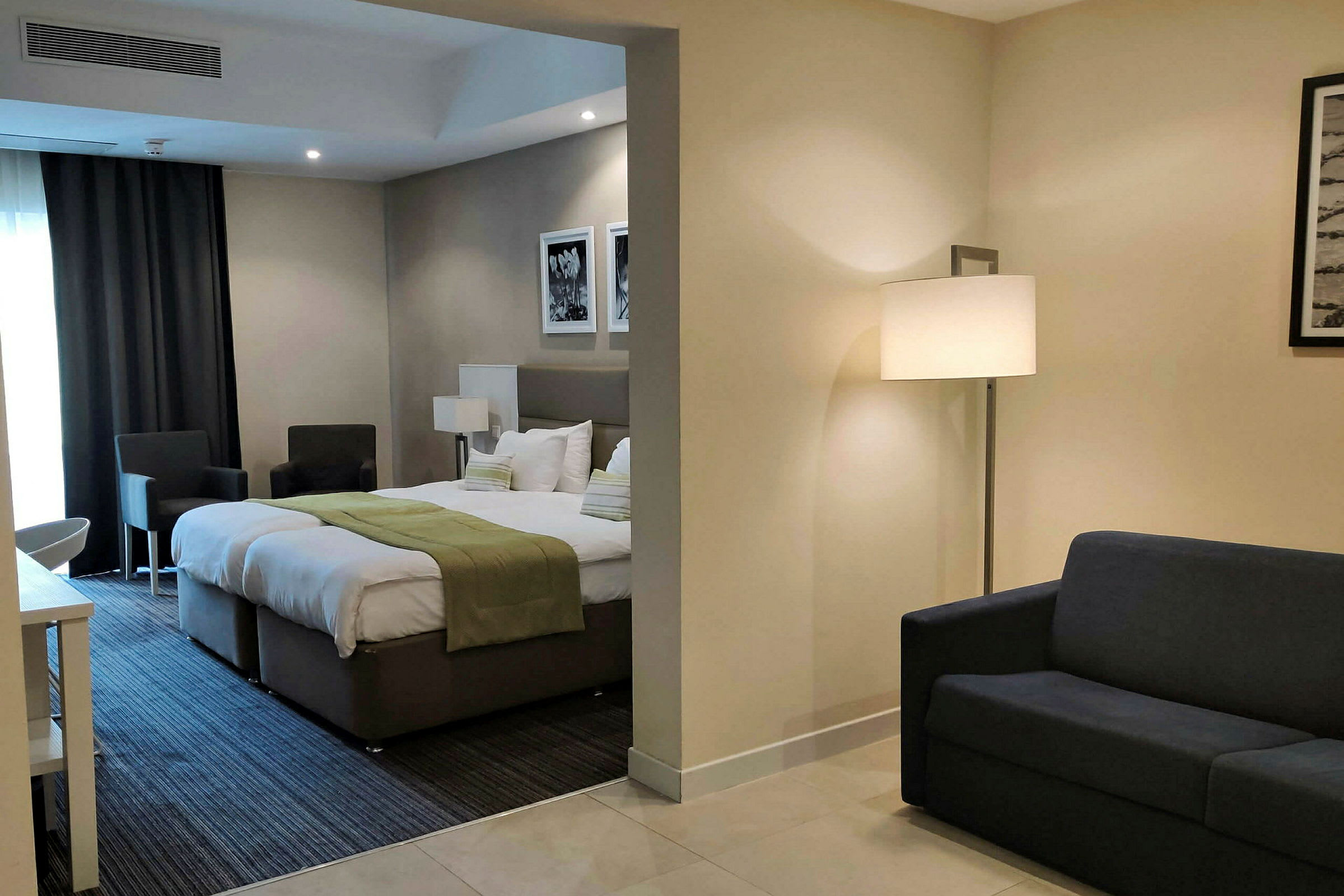 Et hotellrom med en seng, grønt pledd, sittemøbler og en sofa i separat del. (AI-generert tekst)