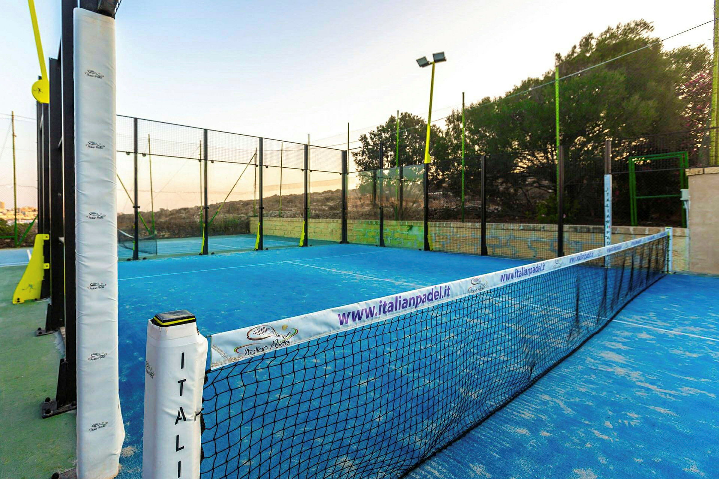 En padelbane med blå markeringer, omgitt av nett og gjerde i en utendørs setting. (AI-generert tekst)