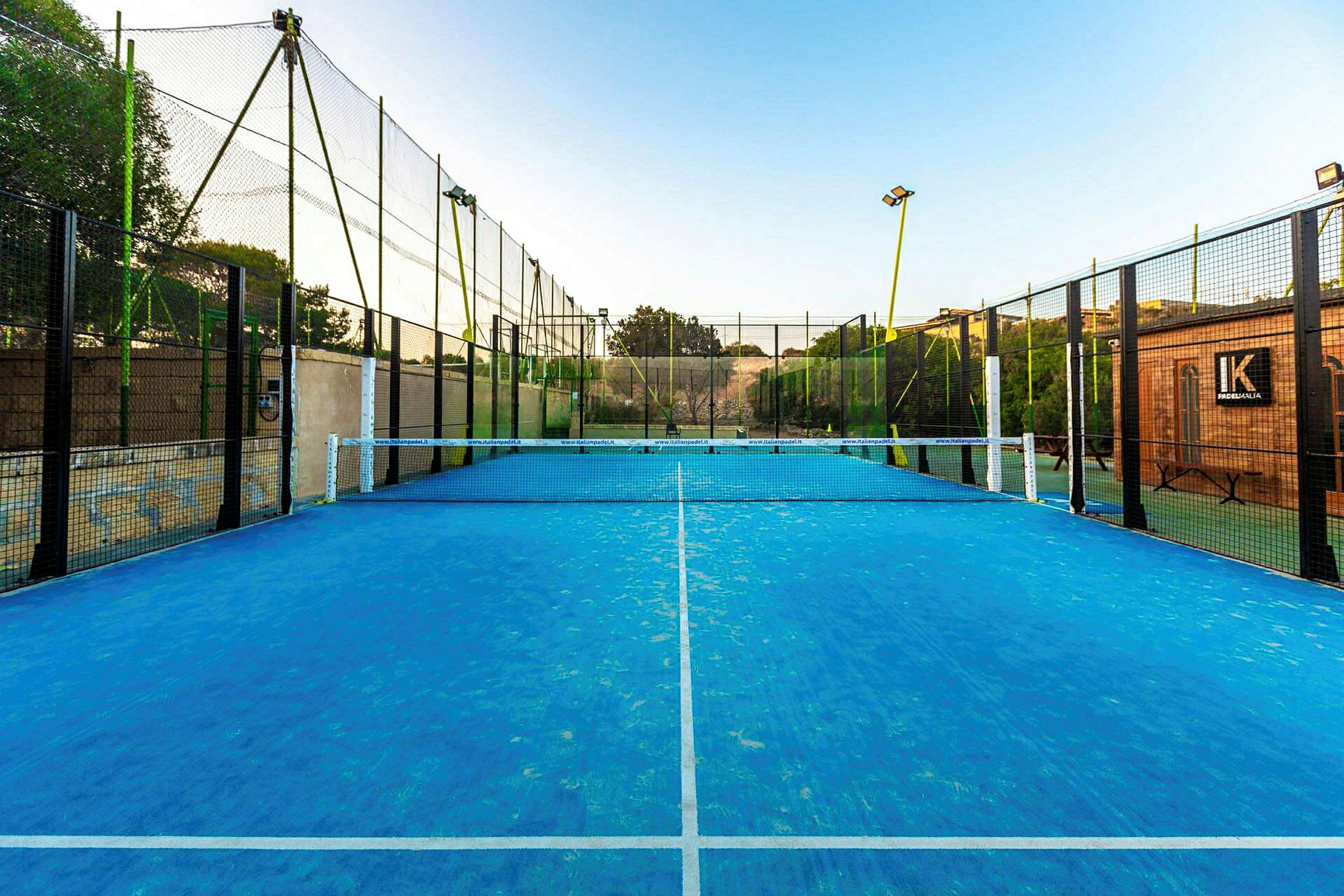 En padelbane med blått underlag og nett i midten, omgitt av gjerder. (AI-generert tekst)