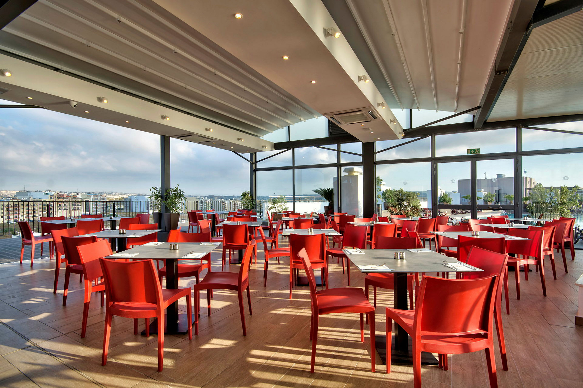 En lys restaurant med røde stoler, trebord og store vinduer med utsikt over en by. (AI-generert tekst)