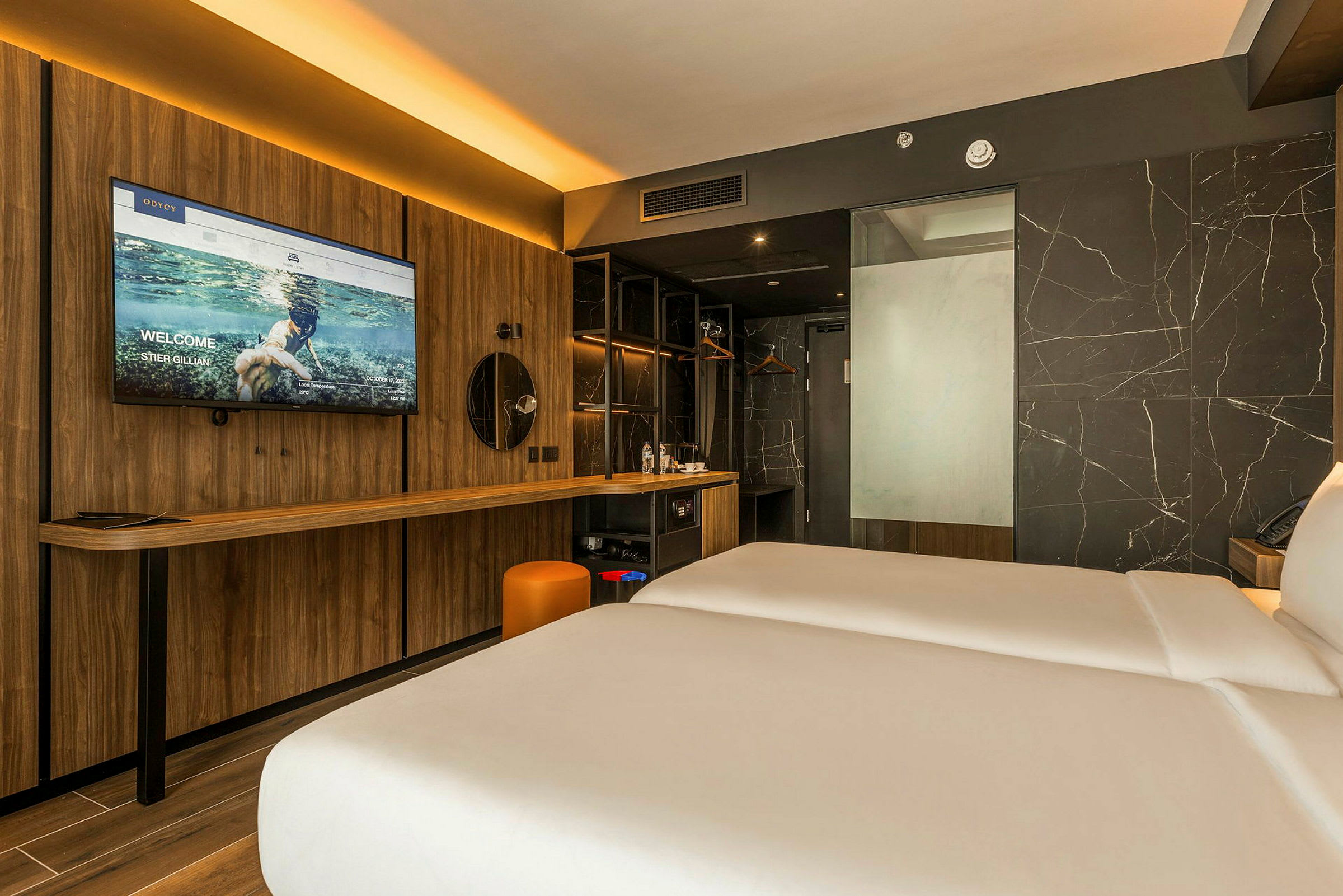 Et moderne hotellrom med to senger, en trevegg med en stor TV og et speil, et skrivebord og en vegg dekorert med marmor. (AI-generert tekst)