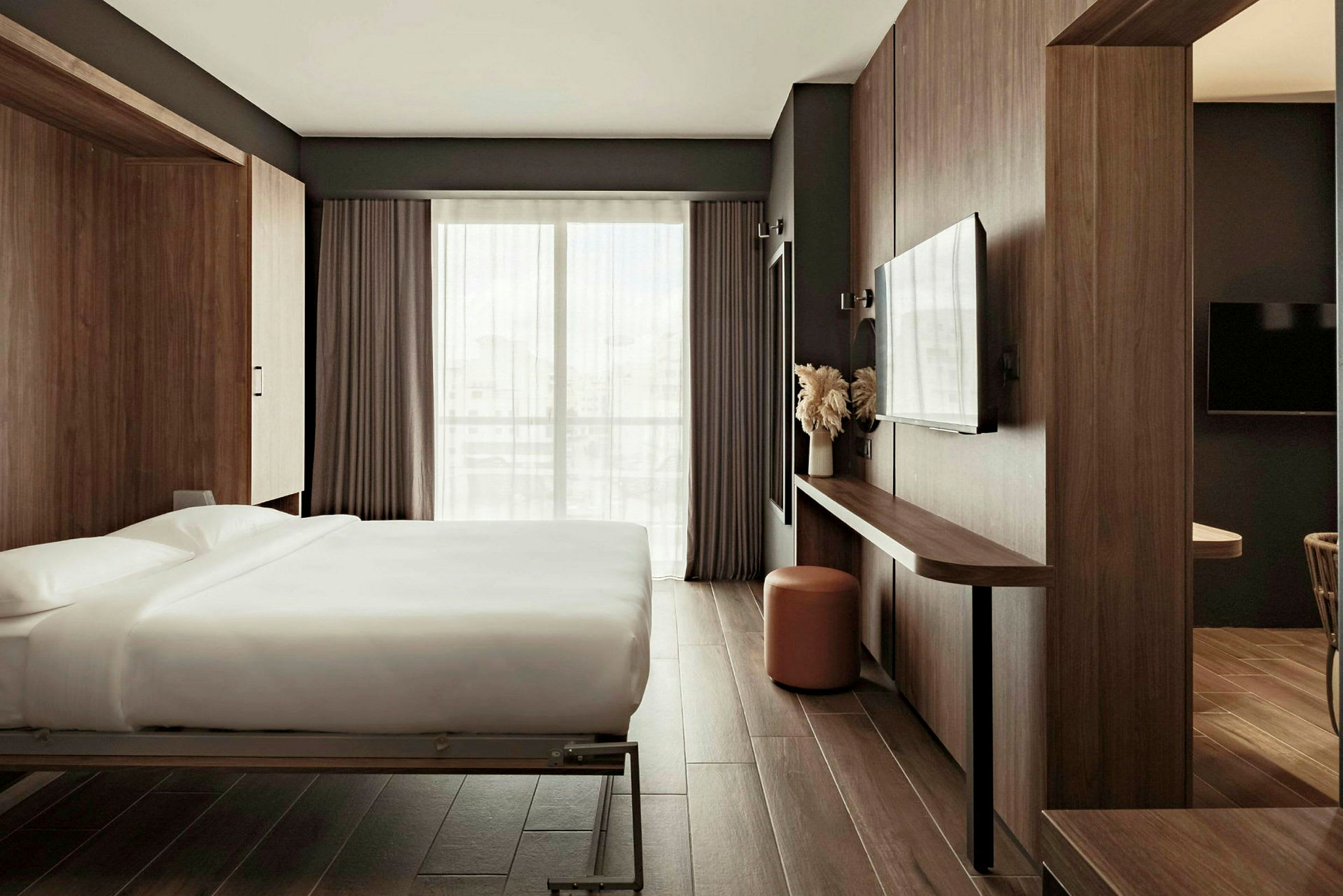Et moderne hotellrom med en dobbeltseng, mørke tregulv og innredning. Rommet har store vinduer med gardiner. (AI-generert tekst)