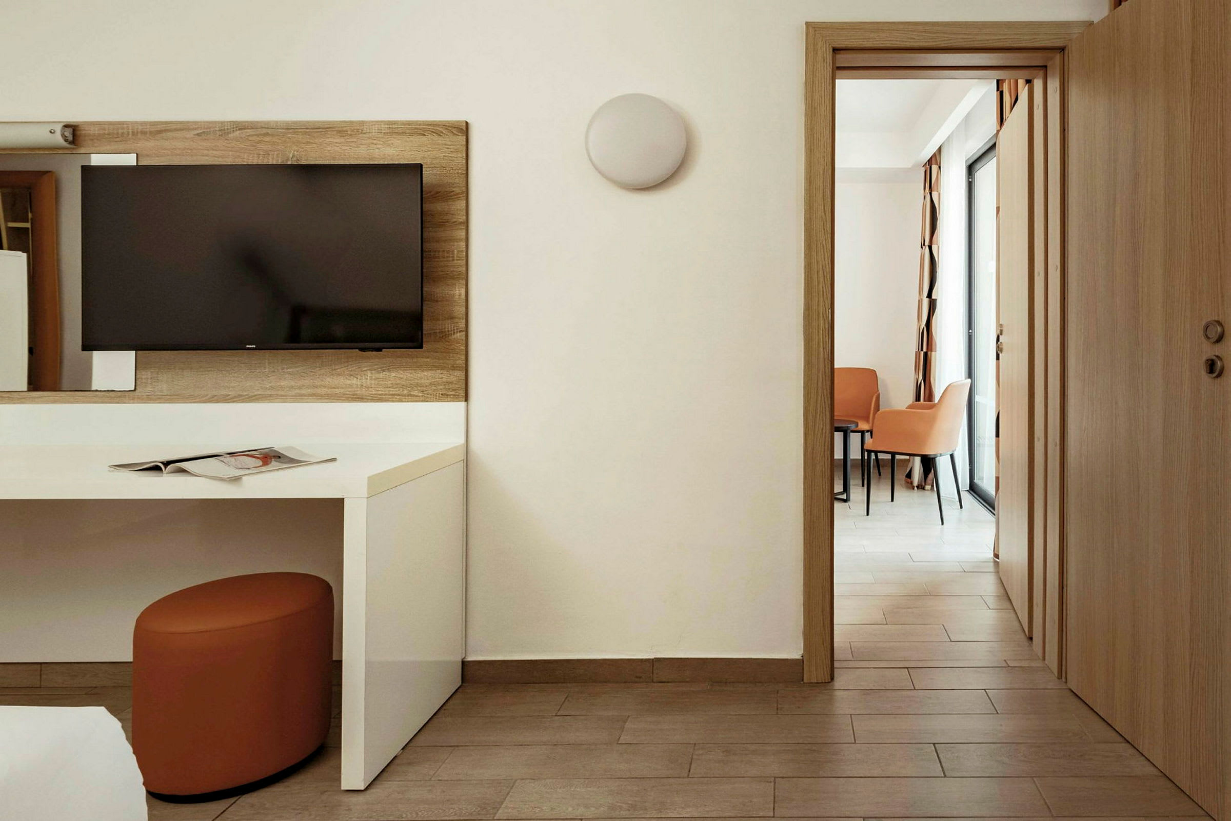 Et hotellrom med en TV plassert på veggen over et hvitt skrivebord. En dør fører til et område med to stoler og et bord. (AI-generert tekst)