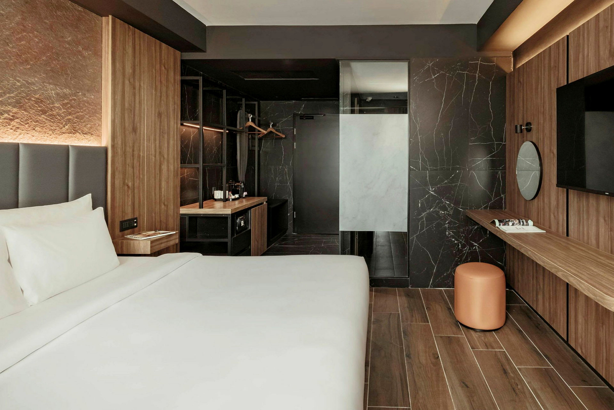 Et moderne hotellrom med en stor seng, trepaneler på veggene og et skrivebord med et rundt speil samt en stol. I bakgrunnen vises et skap og et mørkt bad. (AI-generert tekst)