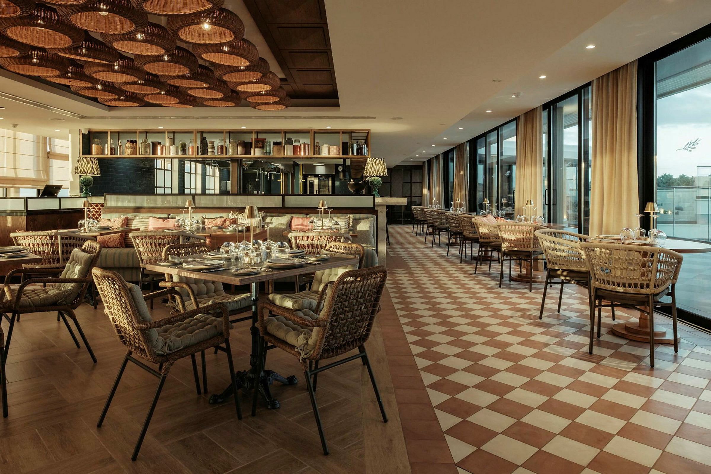 En stilfullt innredet restaurant med bord og rottingstoler. Gulvet har rutemønster og taket er dekorert med flettede lamper. (AI-generert tekst)