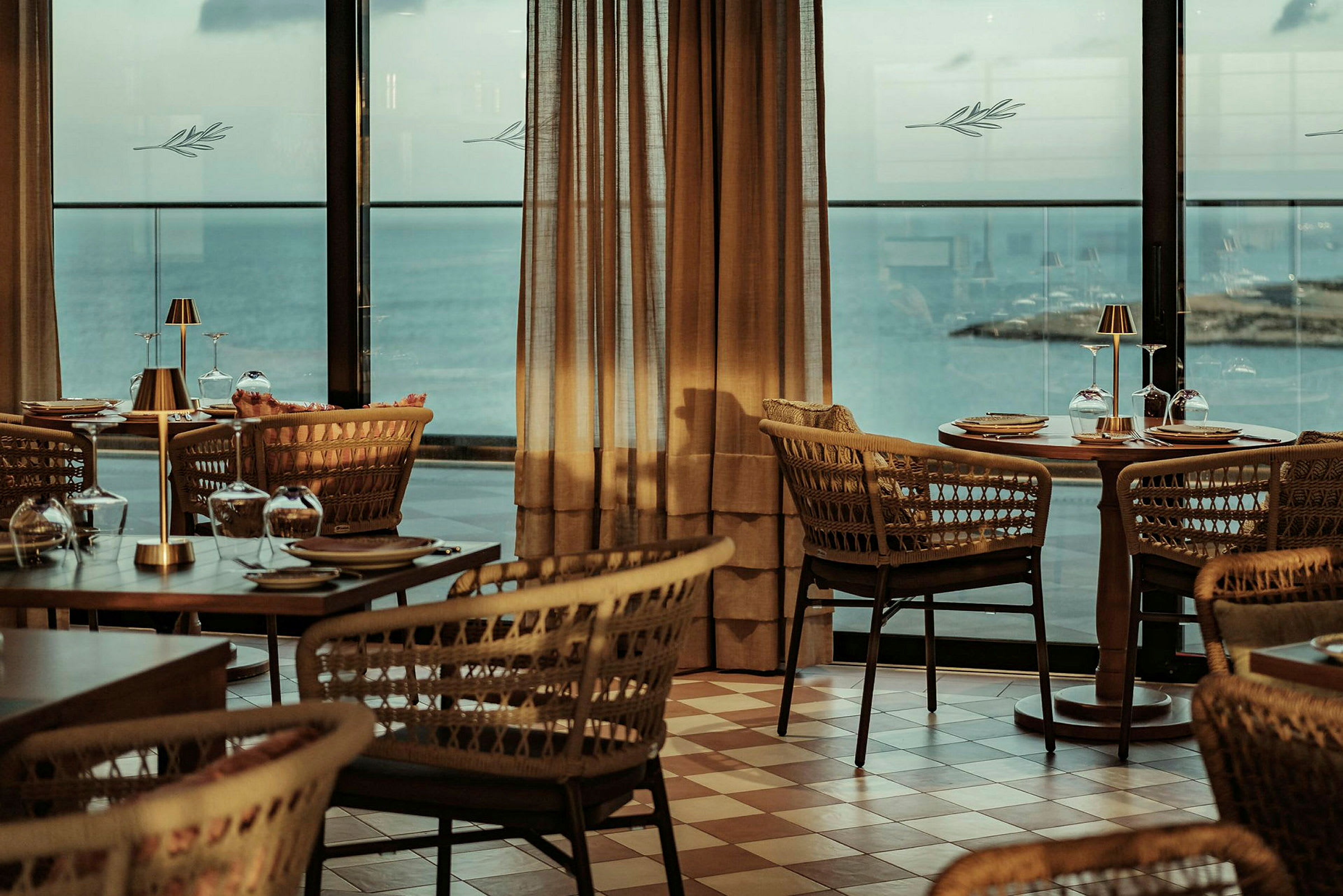 En restaurant med flettede stoler og dekkede trebord med utsikt over havet gjennom store vinduer. (AI-generert tekst)
