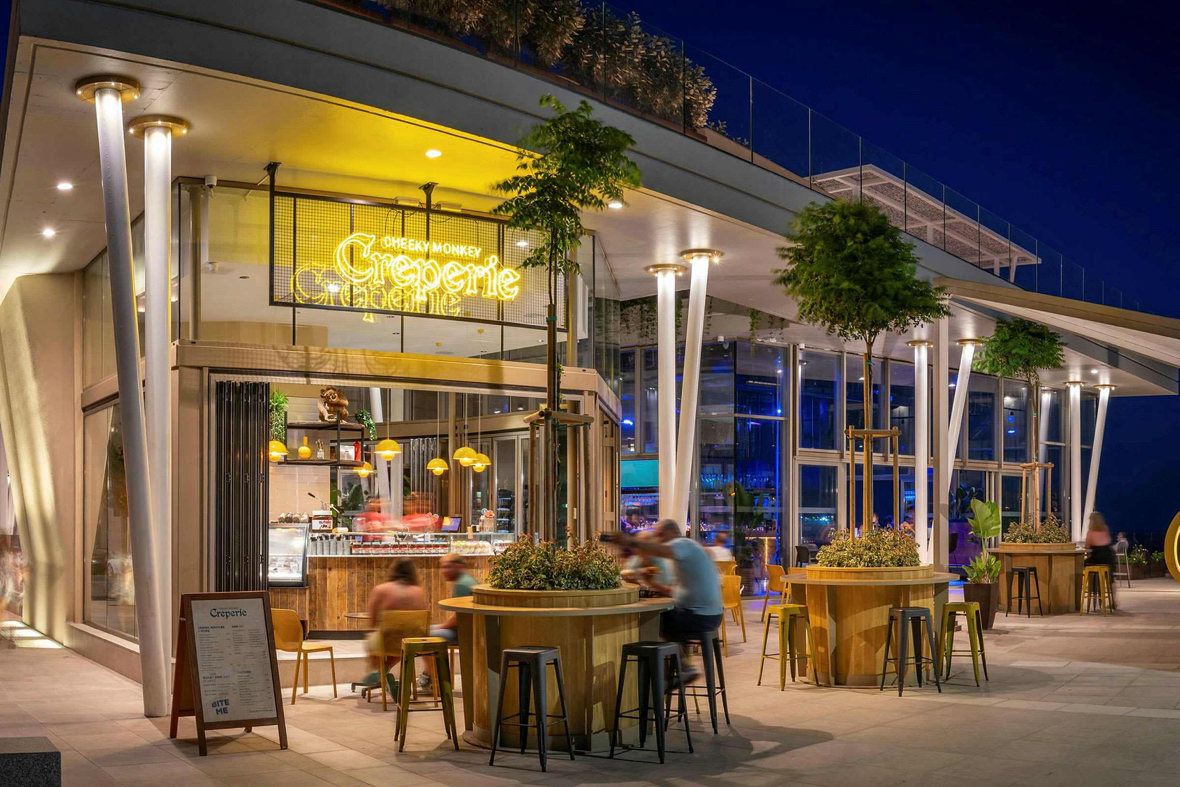 En moderne restaurant eller kafé med opplyst skilting, utendørs sitteplasser og en godt opplyst fasade om kvelden. Trær og plantepynt omgir områdene. (AI-generert tekst)
