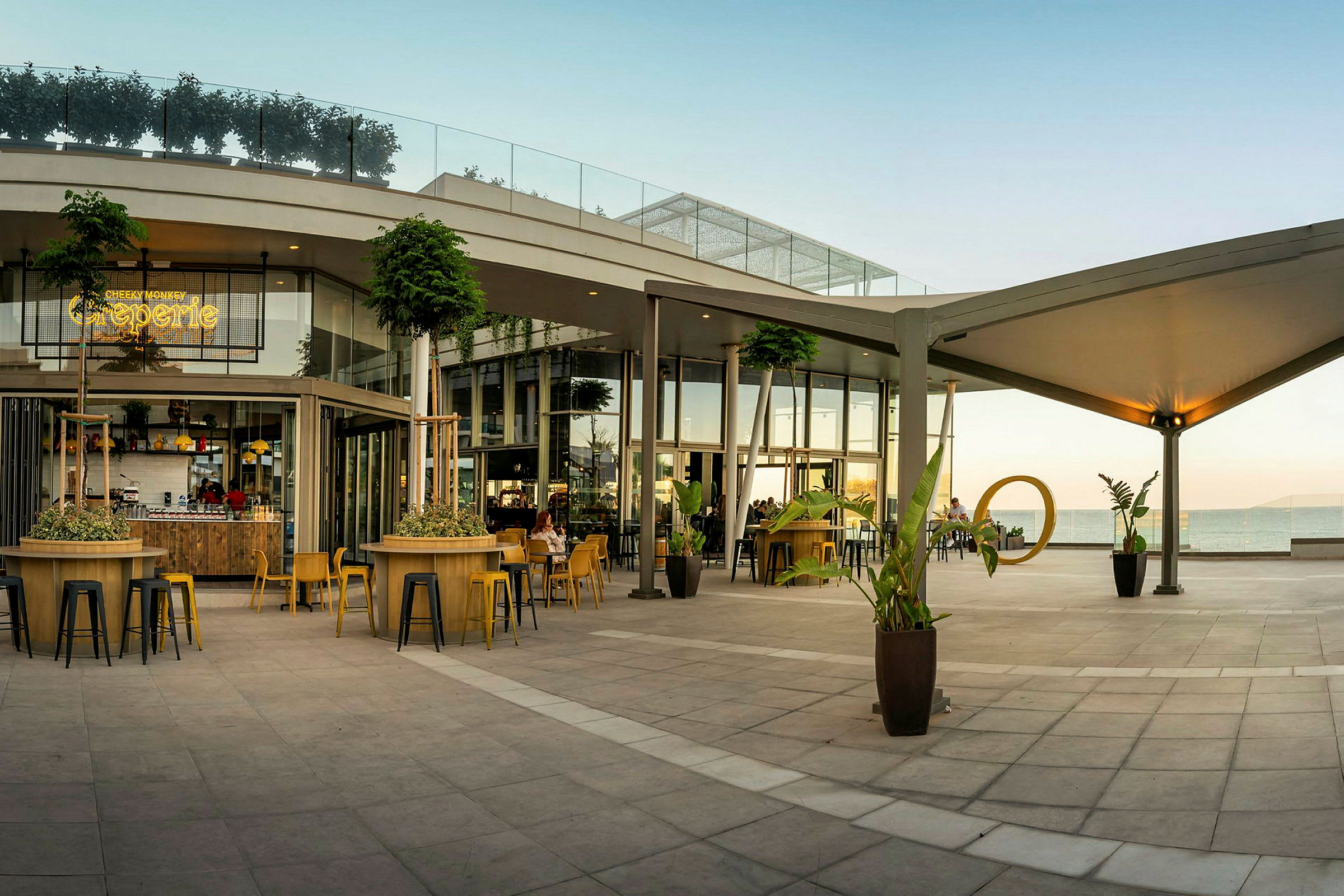 En moderne uteterrasse med sitteplasser og beplantninger ved en kafébygning mot havet. (AI-generert tekst)
