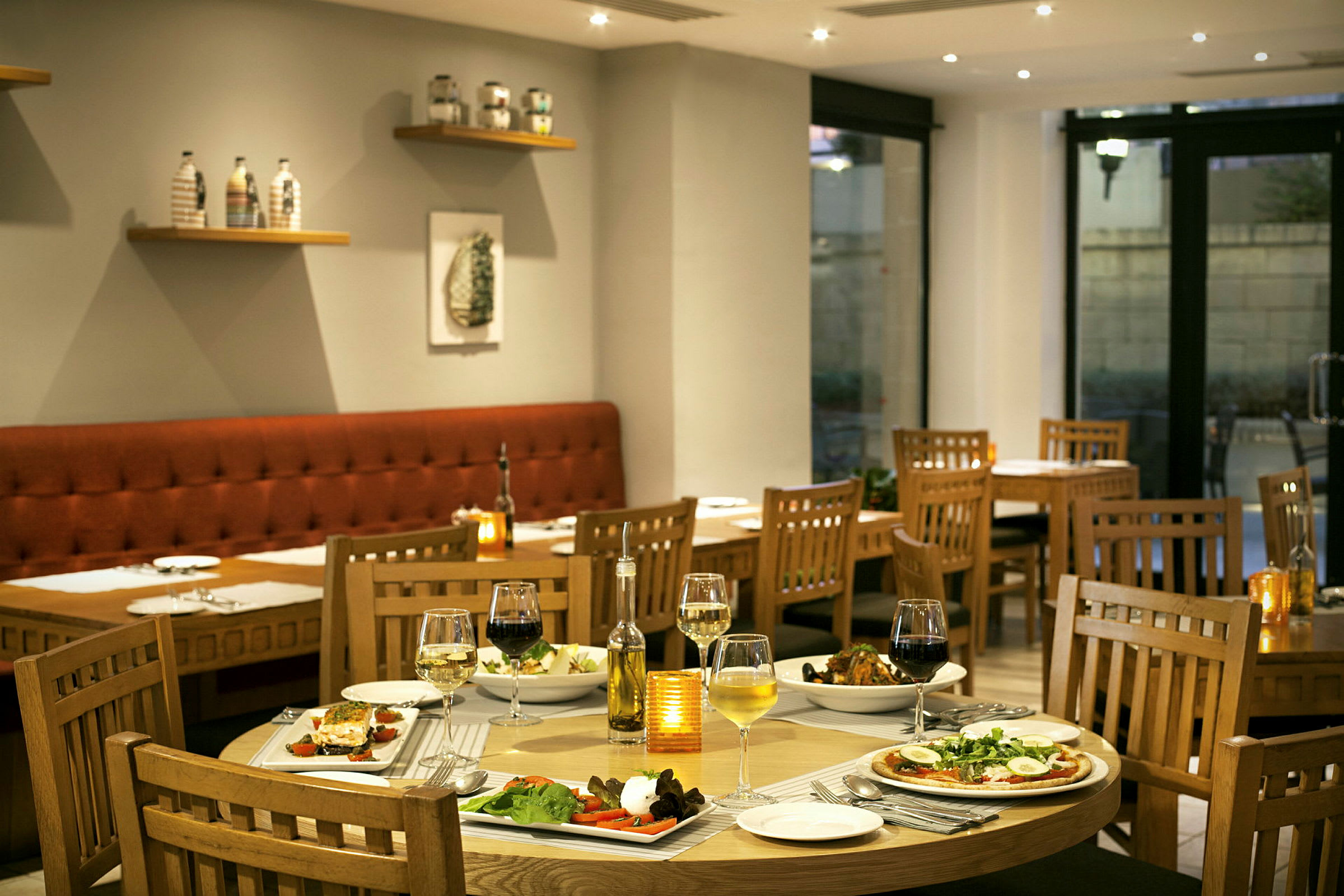 Et smakfullt innredet restaurantområde med dekkede bord og ulike retter. Stoler og bord er av tre, og en komfortabel benkesofa ses langs veggen. (AI-generert tekst)