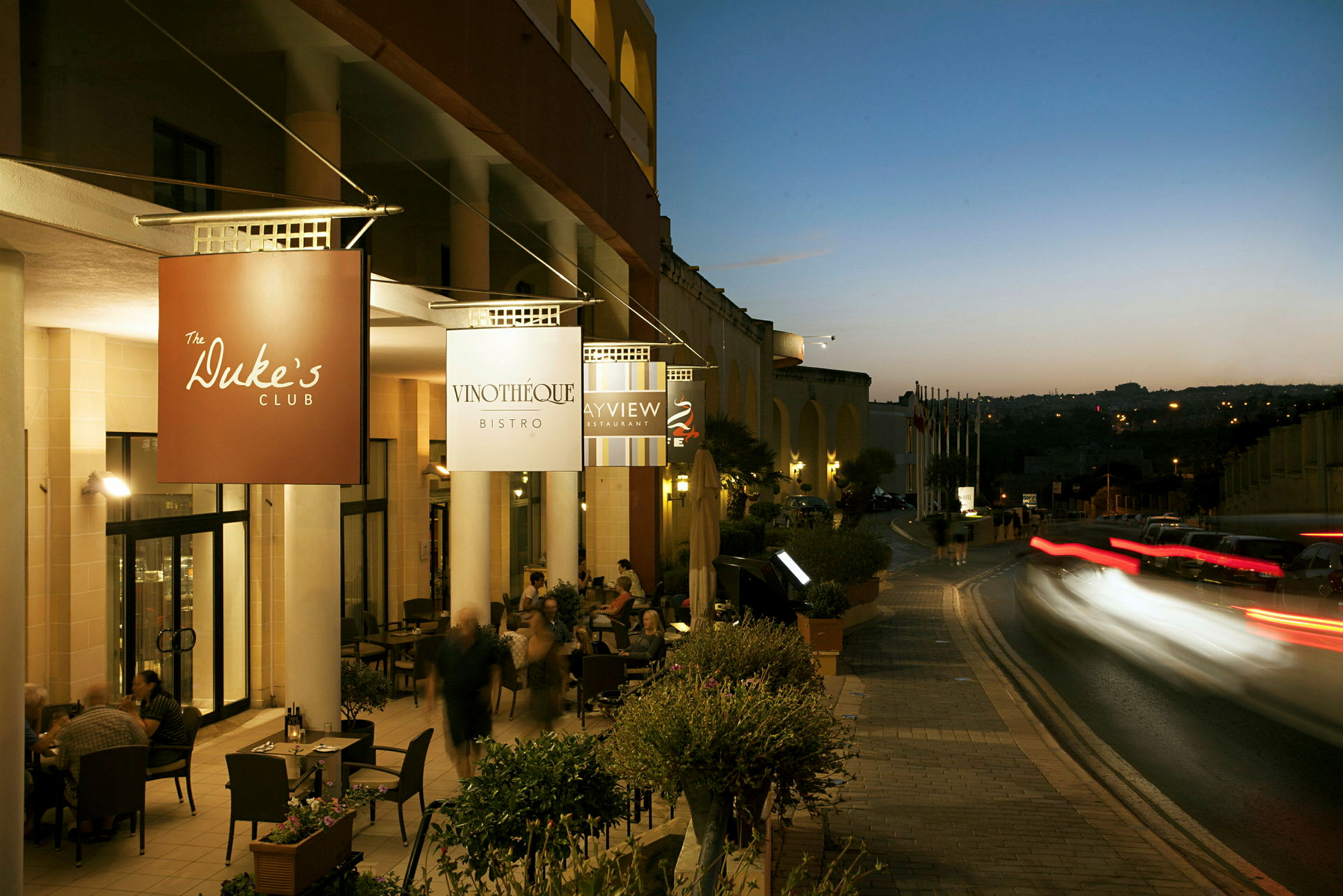 Bilde av en kveldsscene utenfor flere restauranter med opplyste skilt og mennesker, trafikk i bevegelse og nattehimmel i bakgrunnen. (AI-generert tekst)