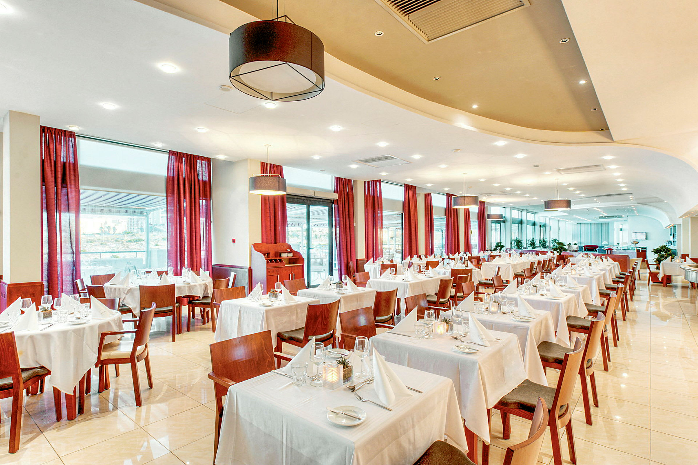 En lys restaurant med bord kledd i hvite duker og røde gardiner ved vinduene. (AI-generert tekst)