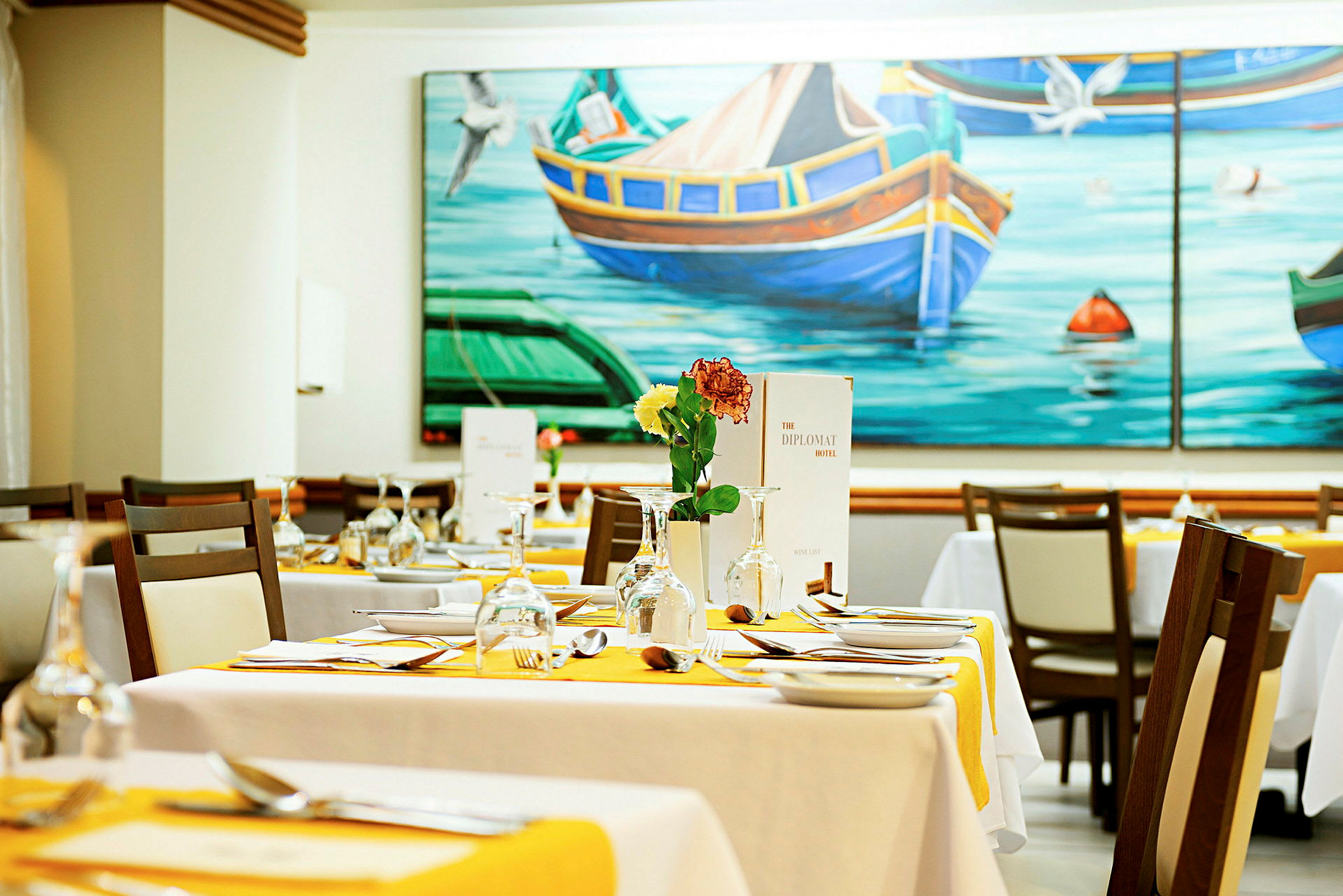 Et restaurantbord med duk og bestikk, dekorert med blomster og glass, med et fargerikt maleri av båter og vann på veggen. (AI-generert tekst)