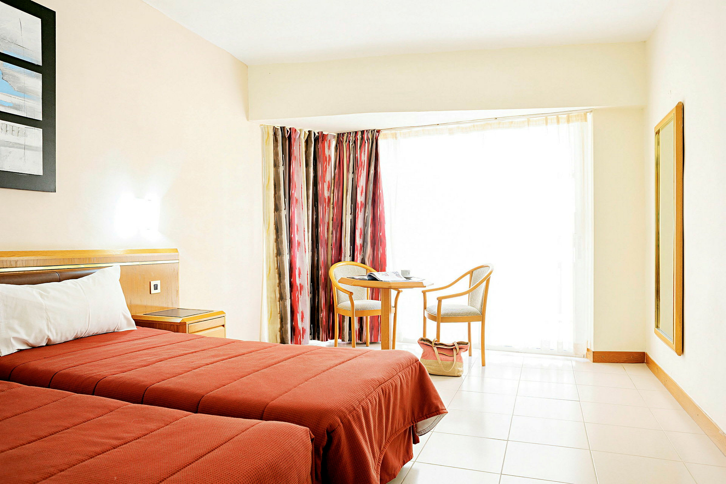 Et lyst hotellrom med to enkeltsenger med røde sengetepper, et lite bord med to stoler ved et vindu med fargerike gardiner, og et speil på veggen. (AI-generert tekst)