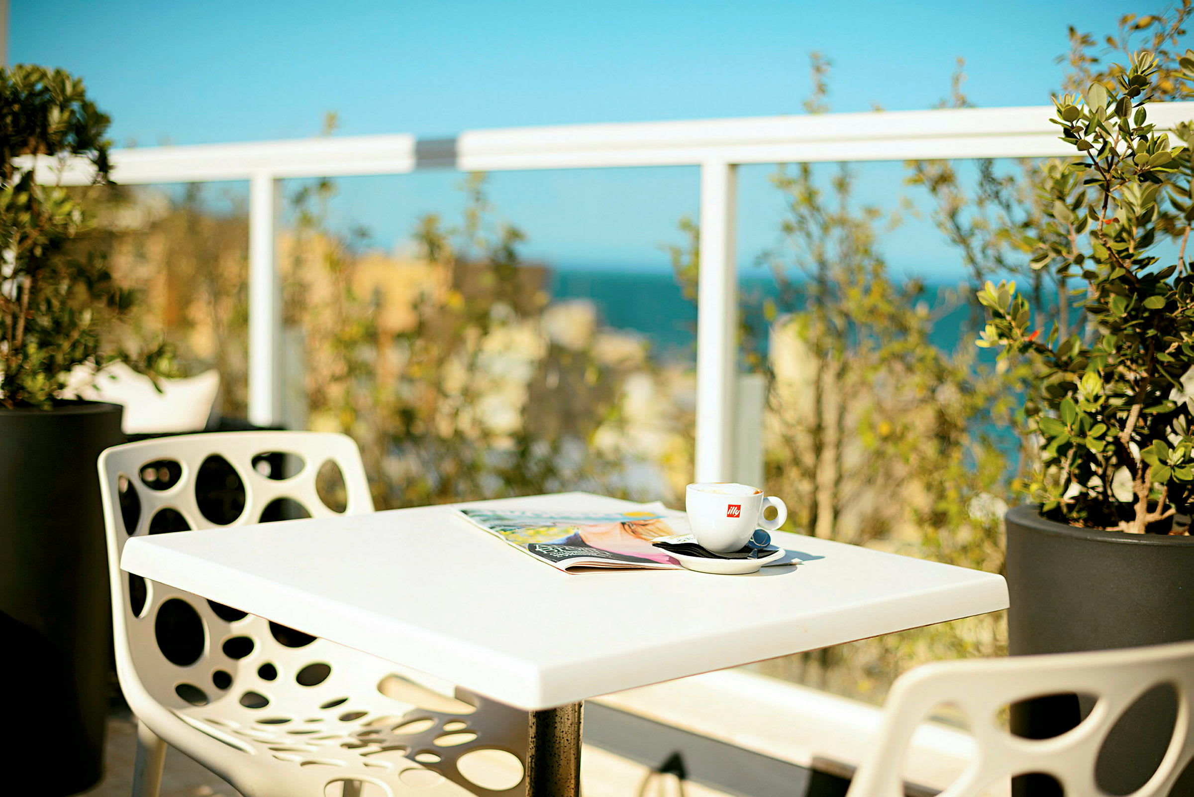 Et bord med en kaffekopp og et blad, omgitt av grøntområder og med utsikt over havet i bakgrunnen. (AI-generert tekst)