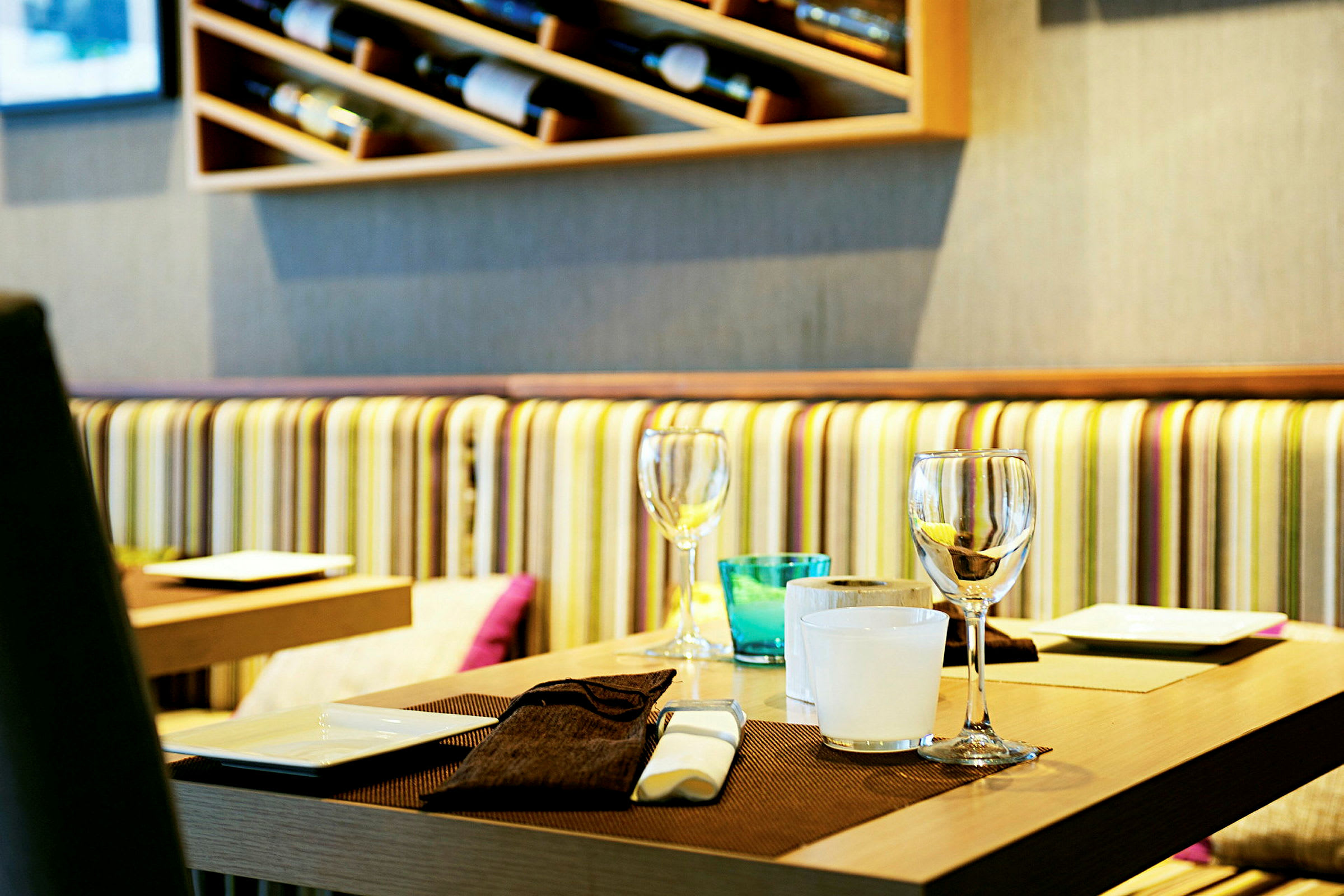 Et restaurantbord med borddekking, inkludert glass, små tallerkener og bordbrikke. Bak bordet vises en vegg med hyller og stripete sofatrekk. (AI-generert tekst)