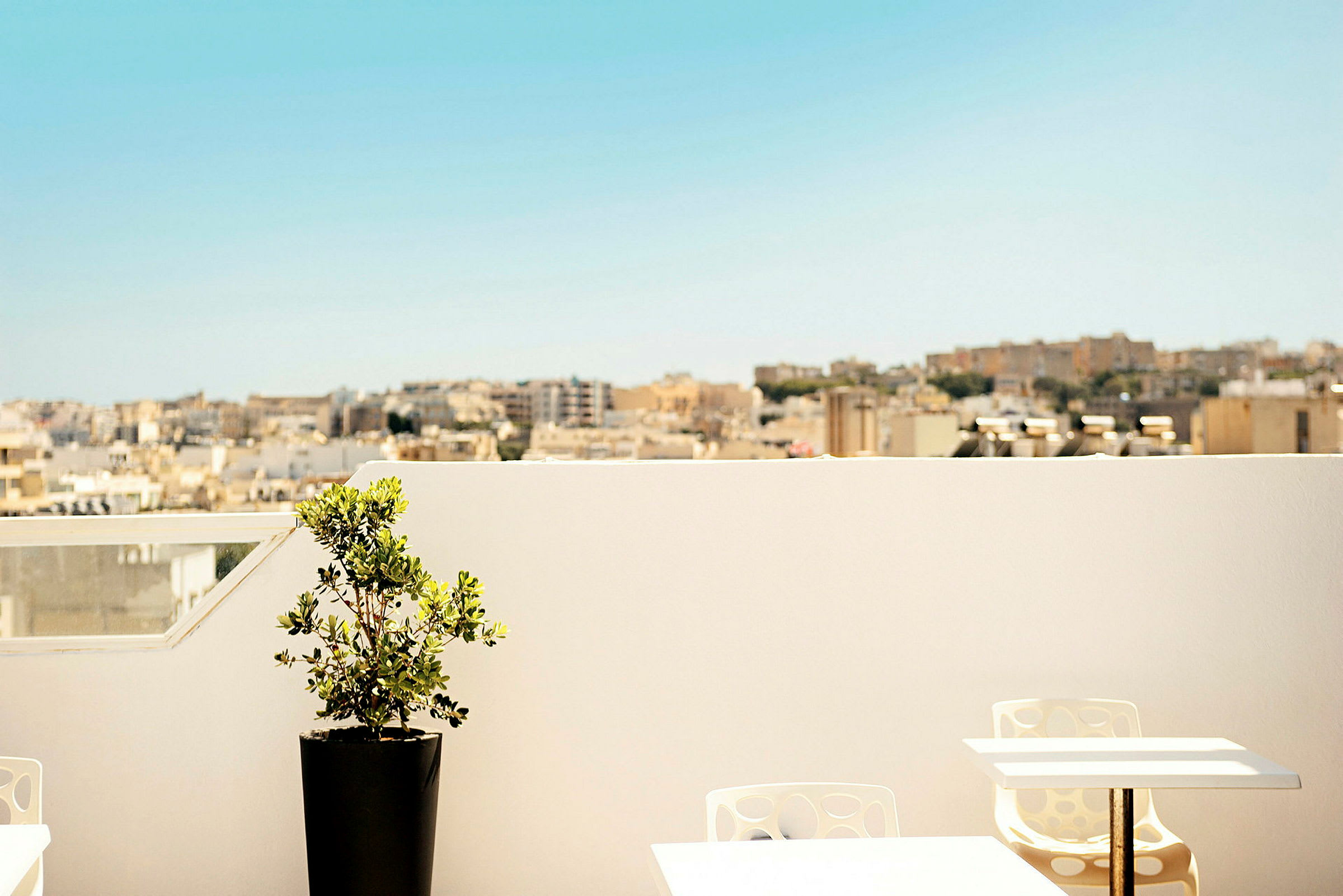 En terrasse med et bord, stoler og en potteplante. I bakgrunnen vises en byutsikt under en klar blå himmel. (AI-generert tekst)