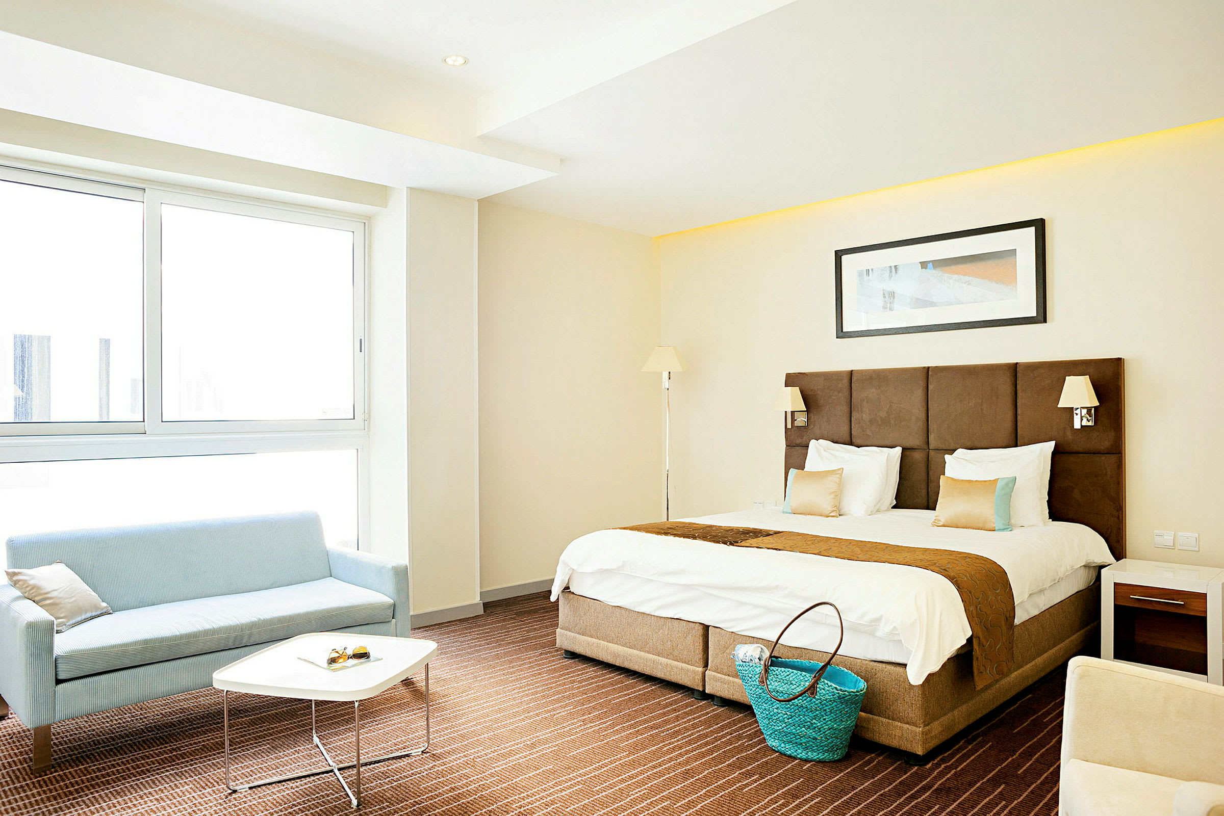 Et lyst og moderne hotellrom med en dobbeltseng, en sofagruppe ved vinduet og et lite bord samt dekorasjoner. (AI-generert tekst)