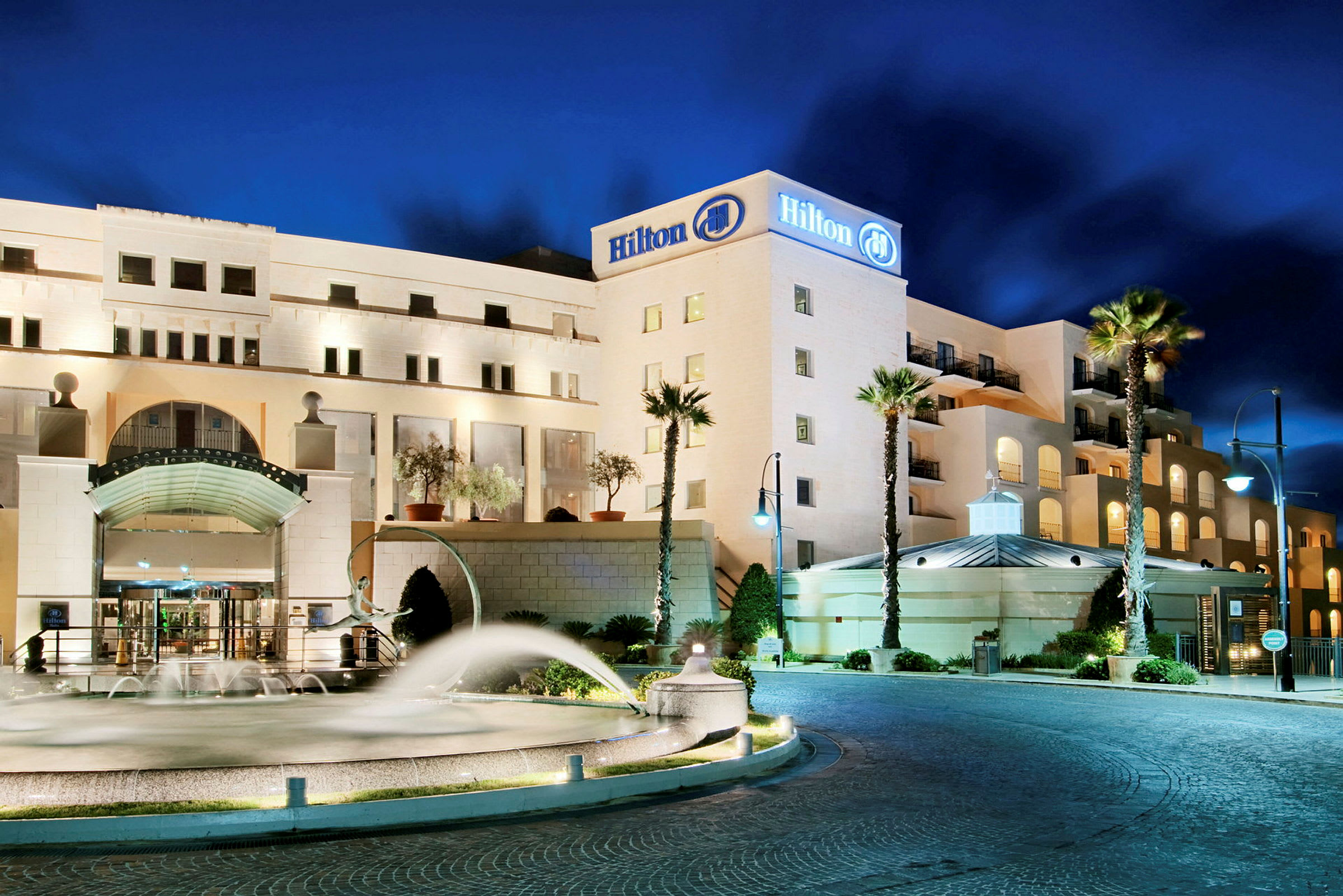 Et hotell med Hilton-logoen på fasaden, opplyst under en nattehimmel. Foran er det en fontene, noen palmer og parkering. (AI-generert tekst)