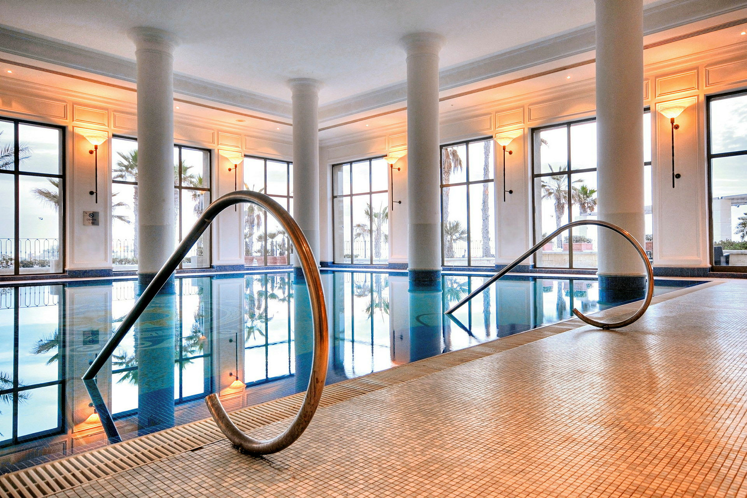 Innendørs basseng med flislagt gulv og store vinduer som vender ut mot palmer utenfor. (AI-generert tekst)