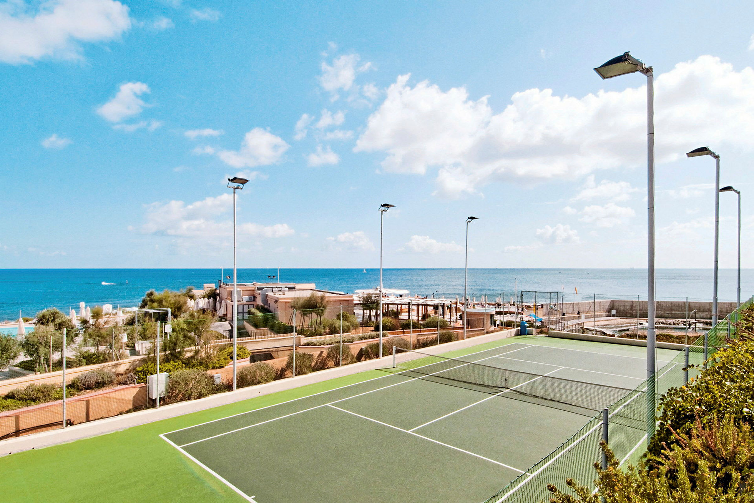 En tennisbane med grønt underlag, omgitt av gjerder og lyktestolper, med havet og bygninger i bakgrunnen. (AI-generert tekst)