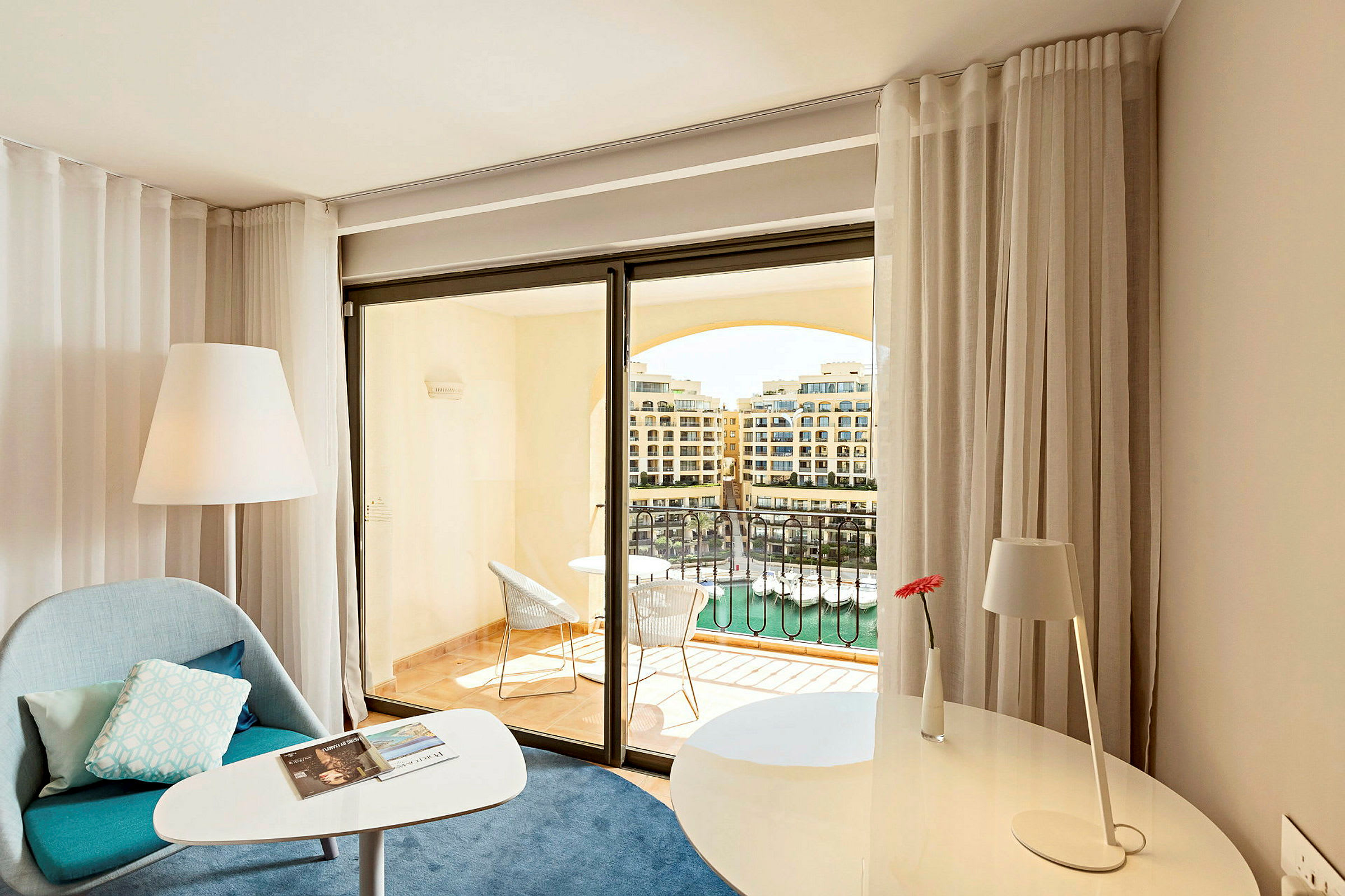 Et lyst hotellrom med sitteområde, skrivebord og glassdør som fører til en balkong med utsikt over en havn. (AI-generert tekst)