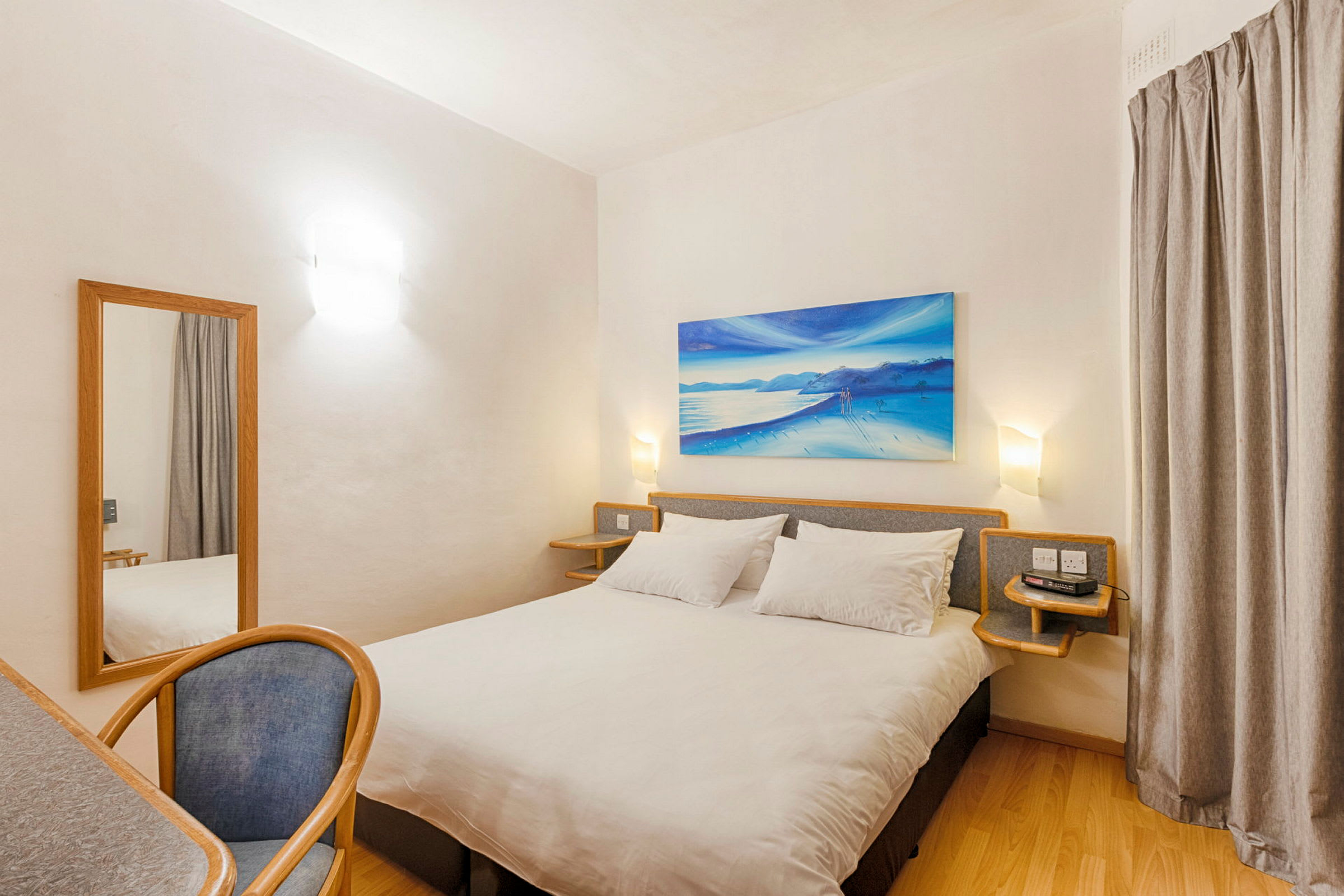 Et hotellrom med en dobbeltseng, et skrivebord med en stol og et speil. Rommet har lyse vegger og en veggdekorasjon med strandmotiv. (AI-generert tekst)
