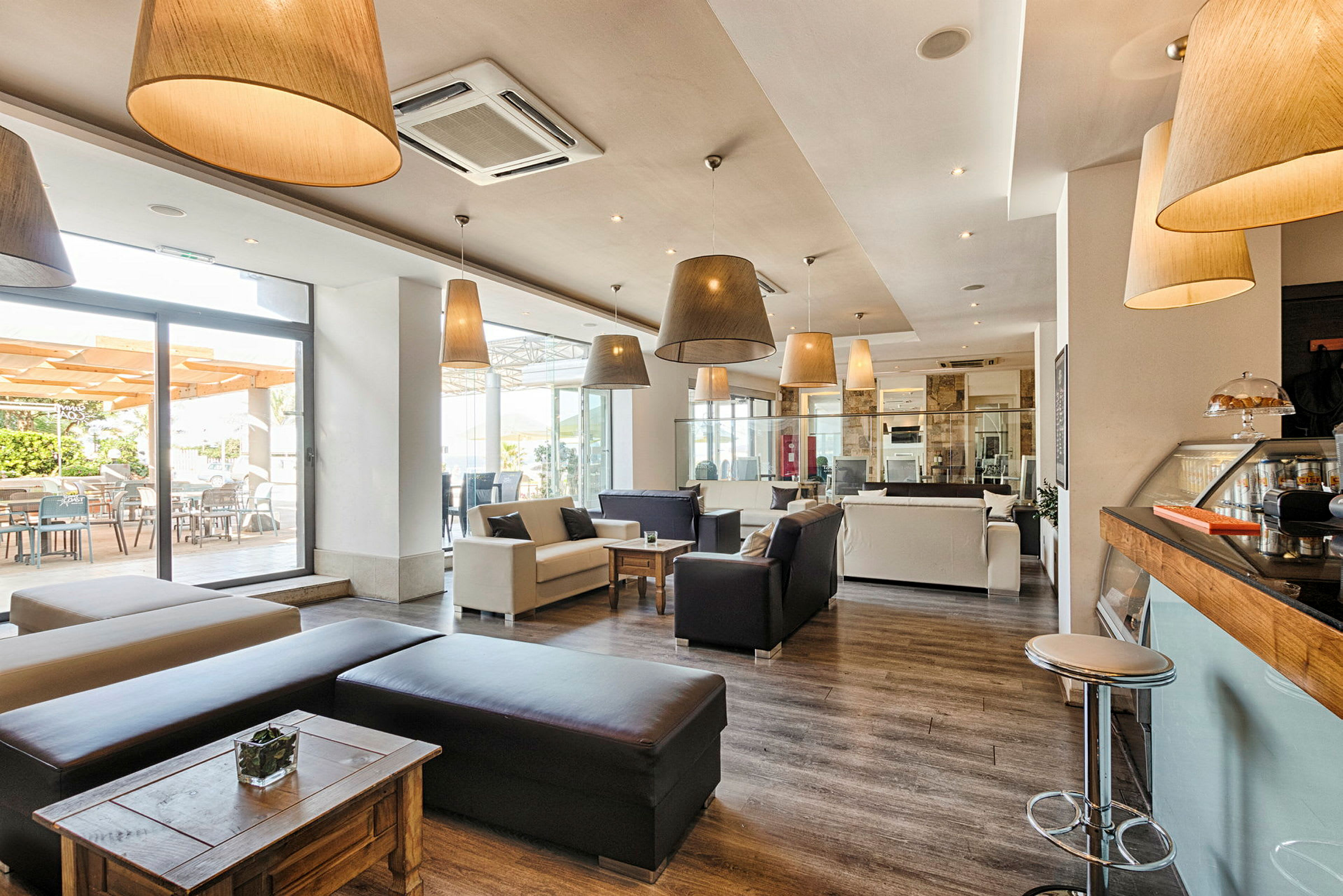 En moderne og romslig kafé eller lounge med sofaer og stoler i lyse farger, lange taklamper og et stort vindusparti som slipper inn dagslys. (AI-generert tekst)