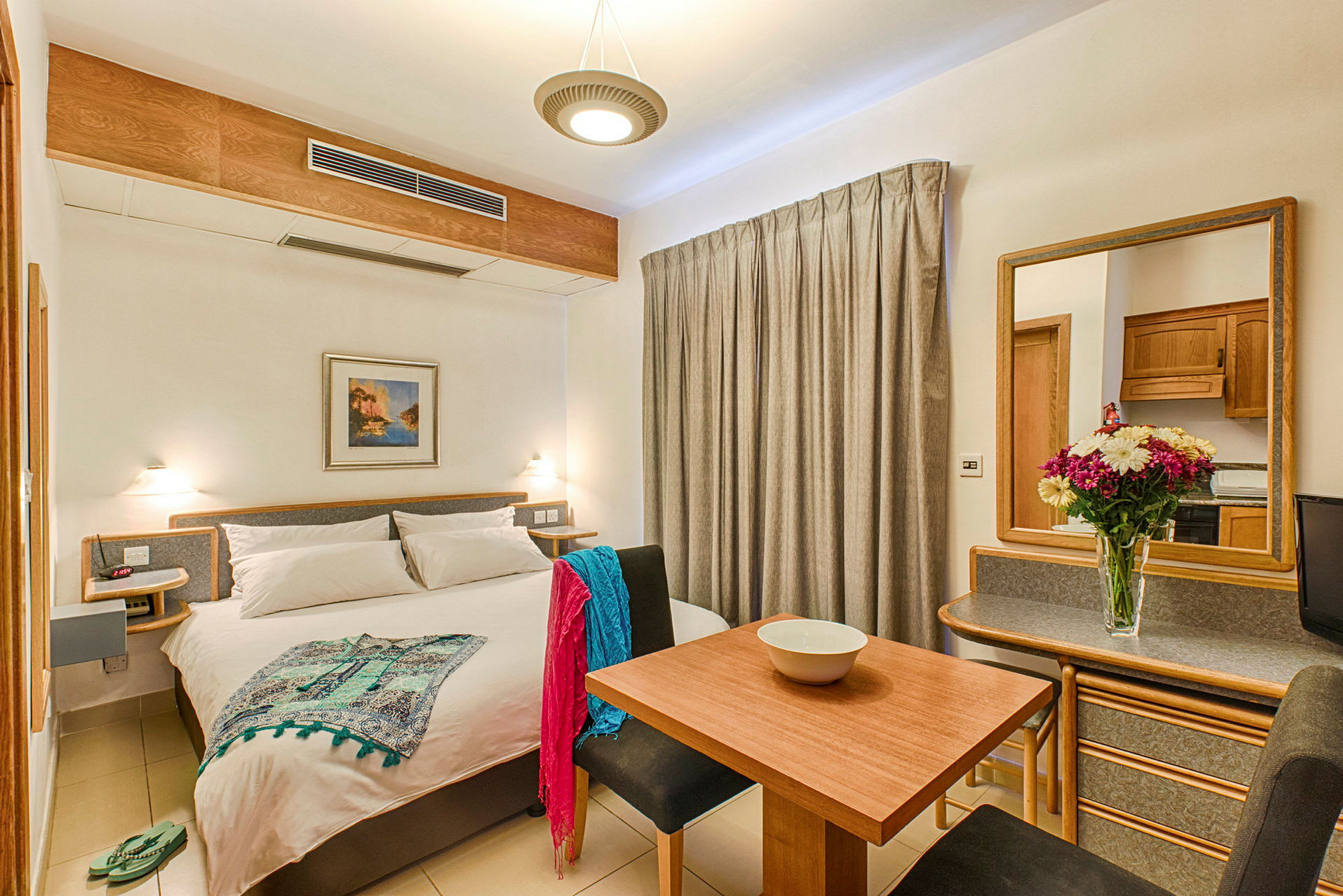 Et lyst hotellrom med en dobbeltseng, spisebord, sitteplasser og et speil. Dekorert med en blomsterpotte på bordet. (AI-generert tekst)