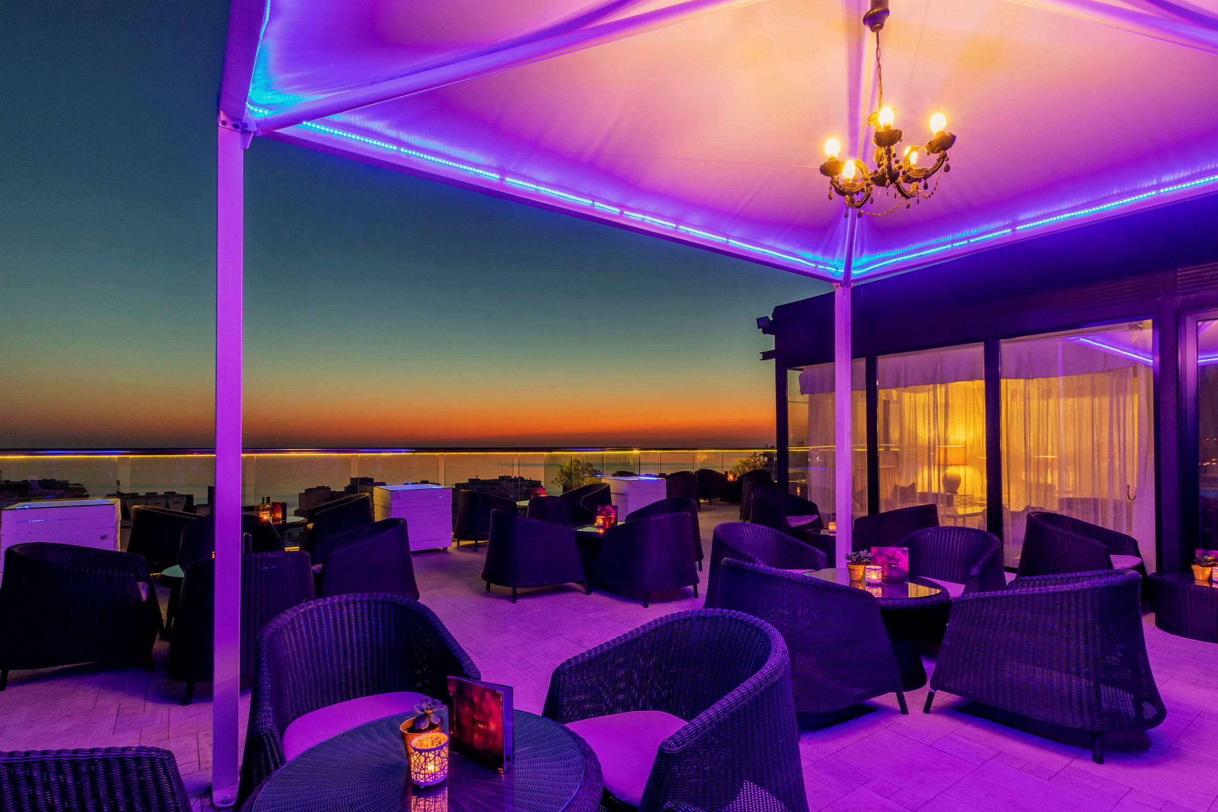En takterrasse med sitteplasser og bord, opplyst med lilla og gule farger mot en solnedgang. (AI-generert tekst)