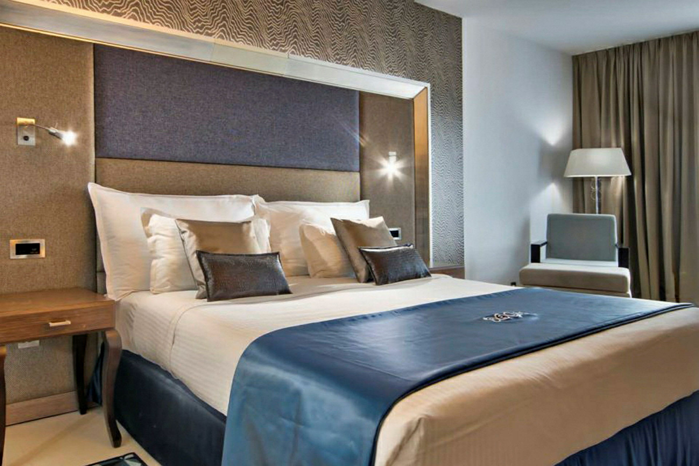 Et hotellrom med moderne interiør som viser en dobbeltseng med hvitt sengetøy og blått sengeteppe, omringet av elegante møbler. (AI-generert tekst)