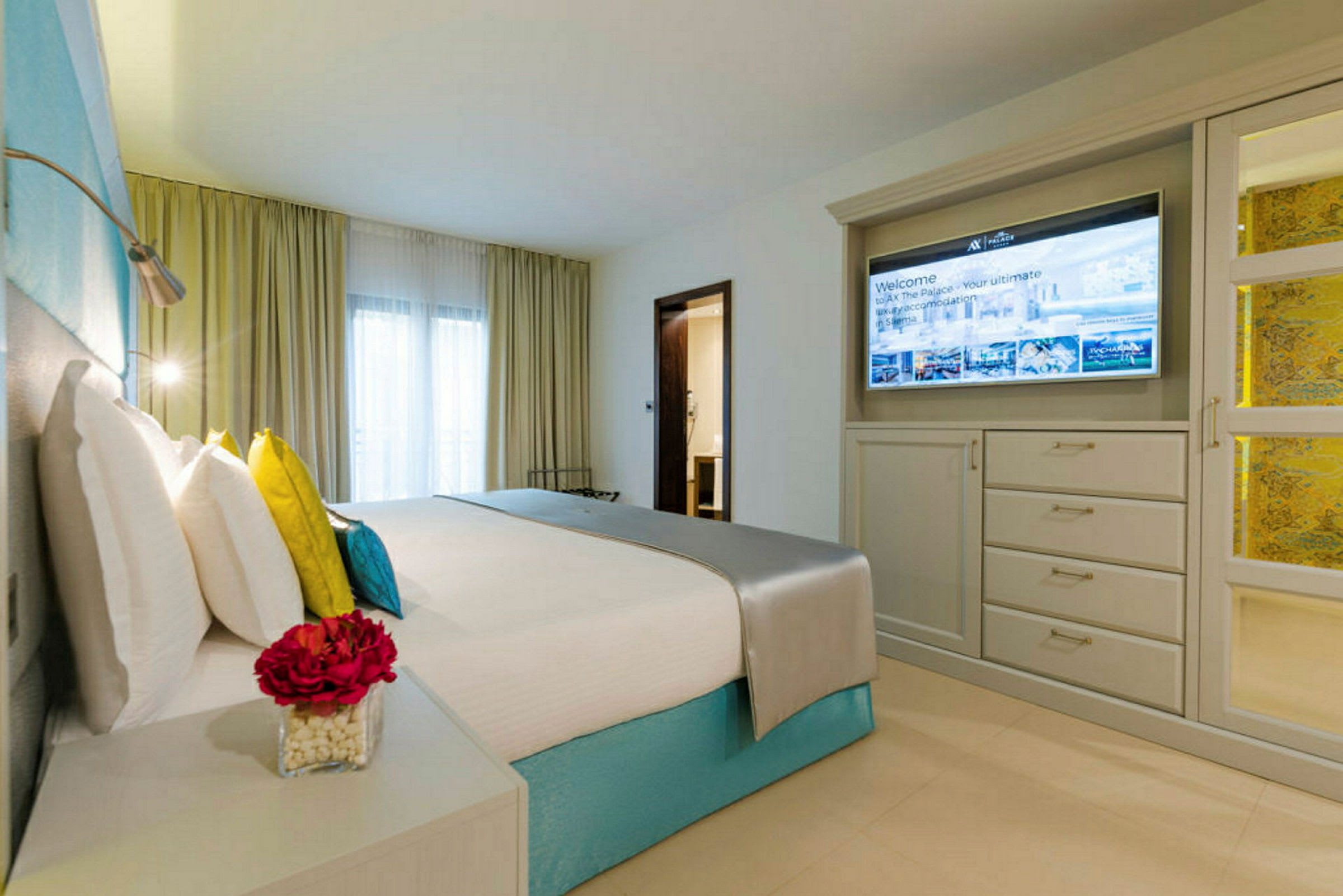 Et moderne hotellrom med en oppredd seng, fargerike puter, et nattbord med en blomsterpotte og en vegg med TV foran et skap. (AI-generert tekst)