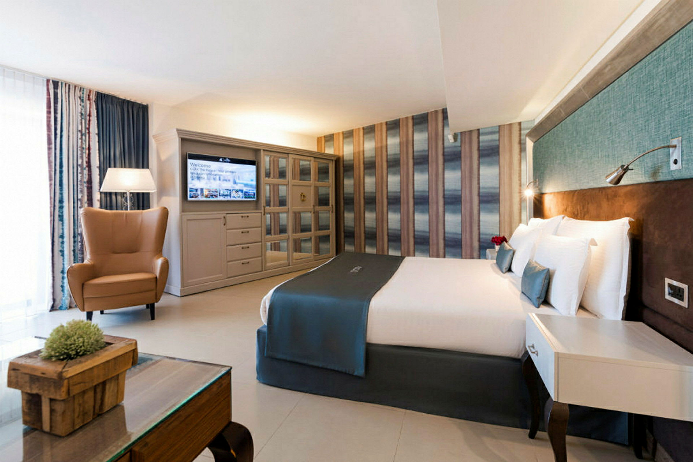 Et hotellrom med en dobbeltseng, en lenestol, et bord og en TV på veggen. (AI-generert tekst)