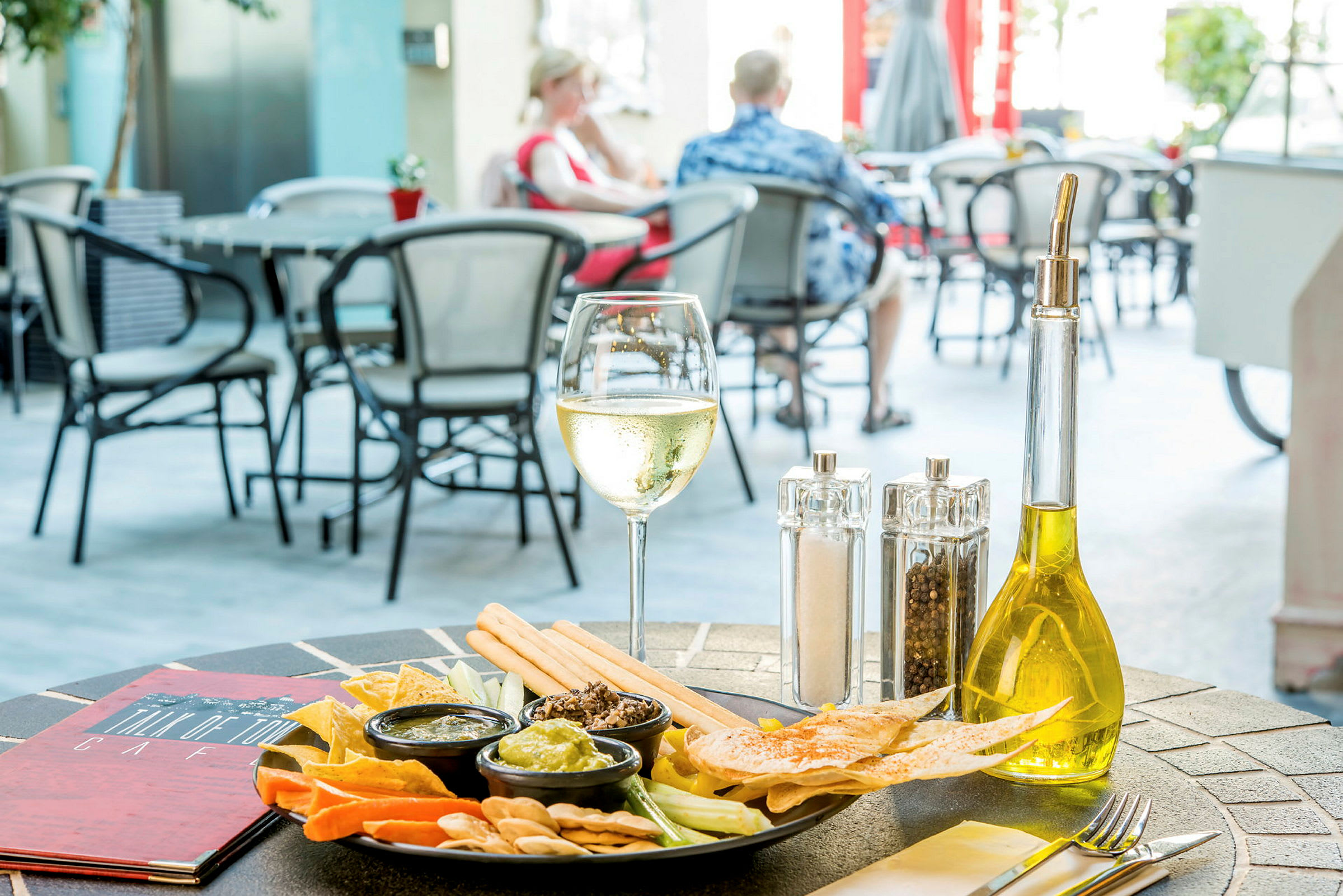 Et bord på en utendørsrestaurant med en tallerken med snacks, små boller med dipp, et glass hvitvin og en flaske olivenolje. (AI-generert tekst)