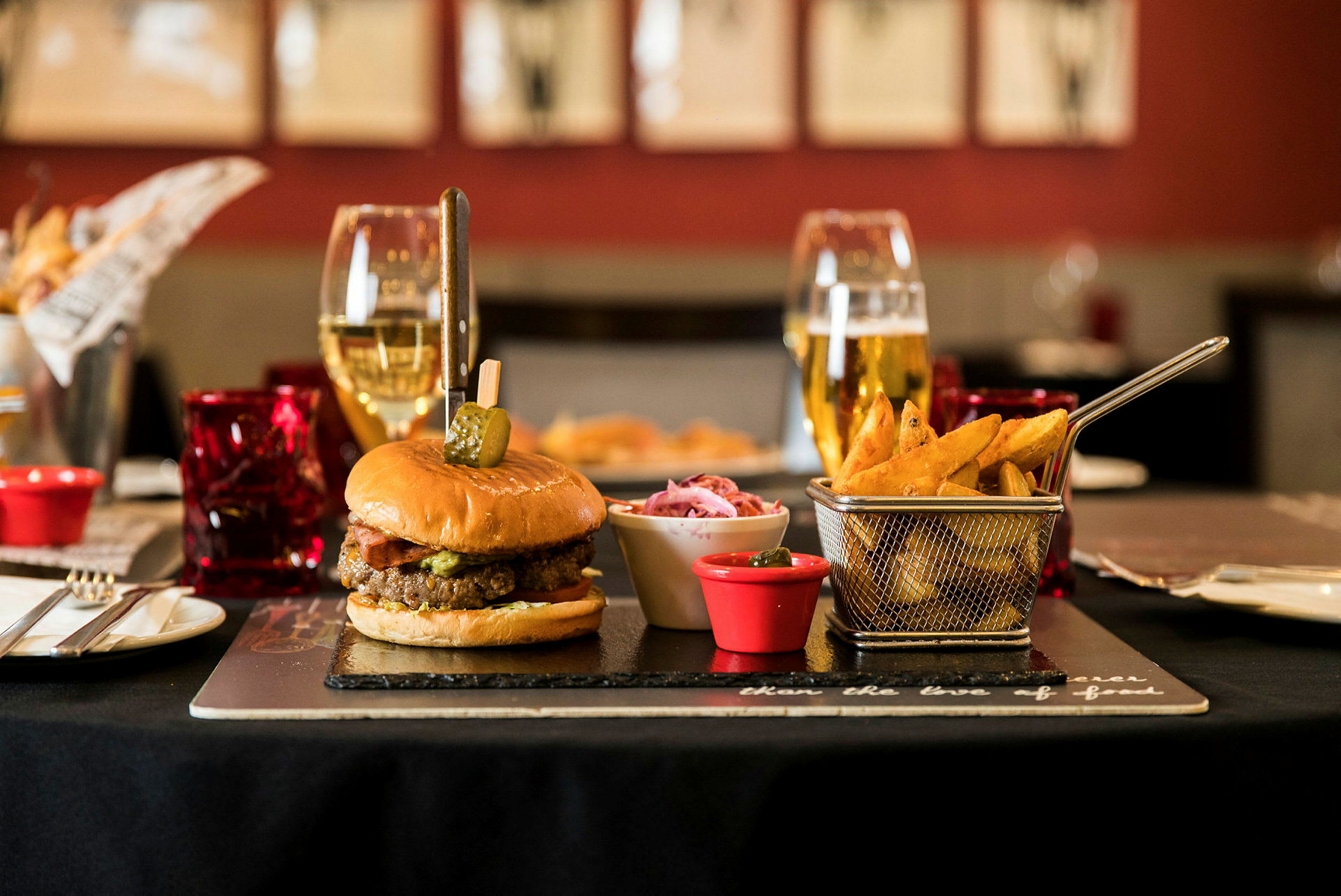 En hamburger med pommes frites og tilbehør på et bord, omgitt av glass og bestikk. (AI-generert tekst)