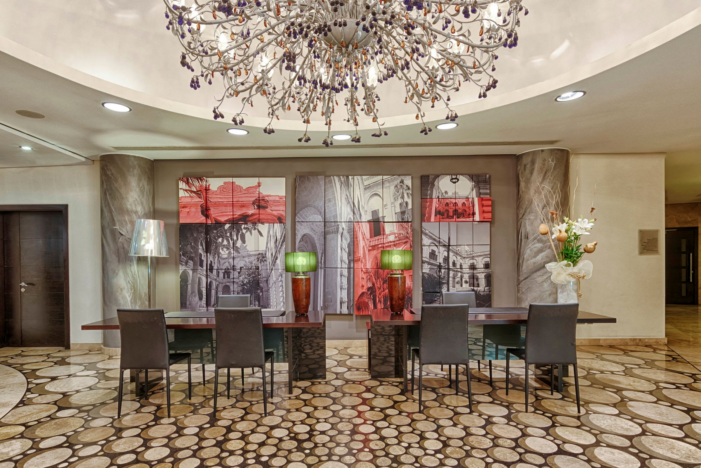 En elegant hotellobby med en stor dekorativ taklampe som henger over flere skrivebord med stoler, malerier på veggen bak, og vakre dekorasjoner som blomster og bordlamper. (AI-generert tekst)