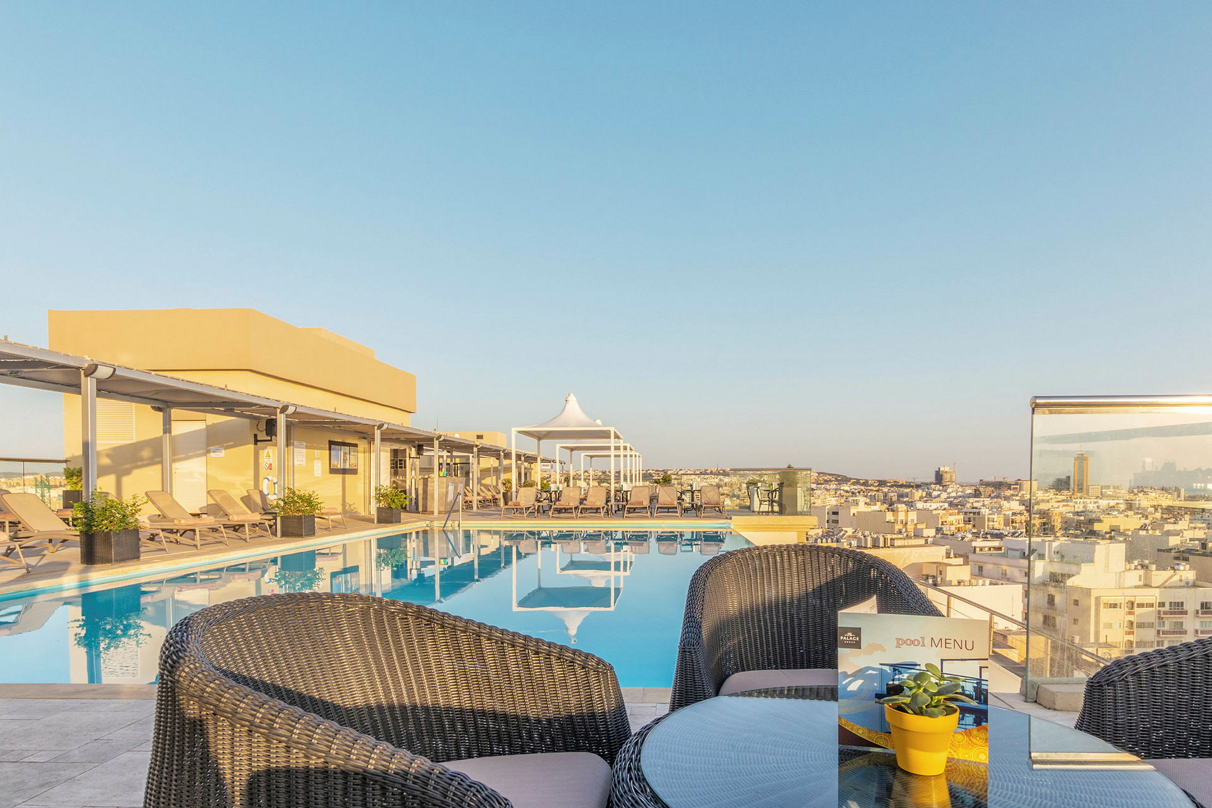 Bilde av et basseng på en hotellterrasse med utsikt over byen, omgitt av solsenger og et mindre bord med stoler. (AI-generert tekst)
