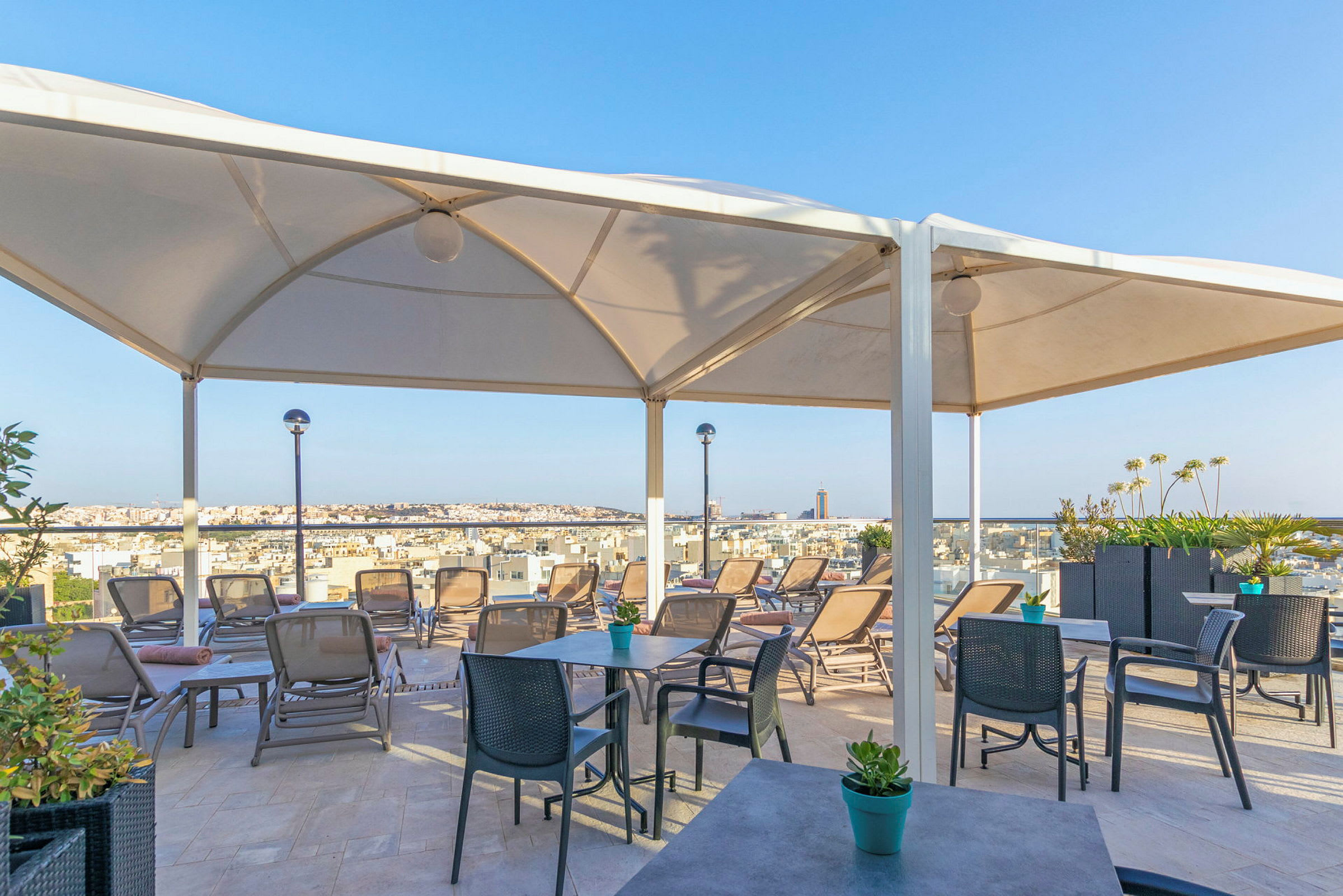 Bilde av en terrasse med solstoler og bord under et stort hvitt tak, med utsikt over en by. (AI-generert tekst)