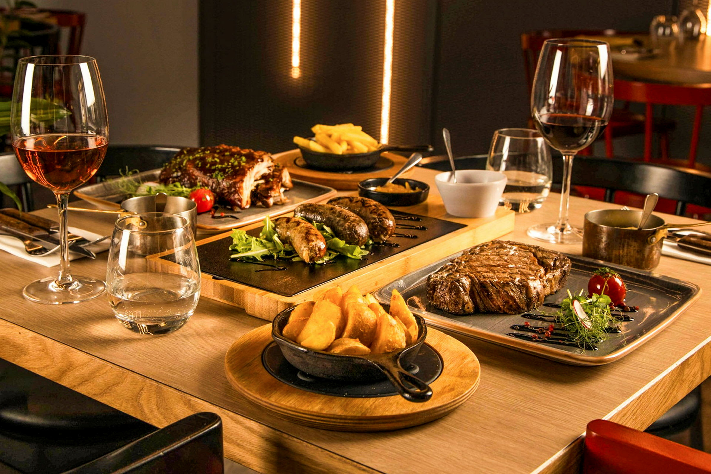 Et bord er dekket med ulike retter, inkludert biff, pølser, ribbe og pommes frites, sammen med vinglass. (AI-generert tekst)