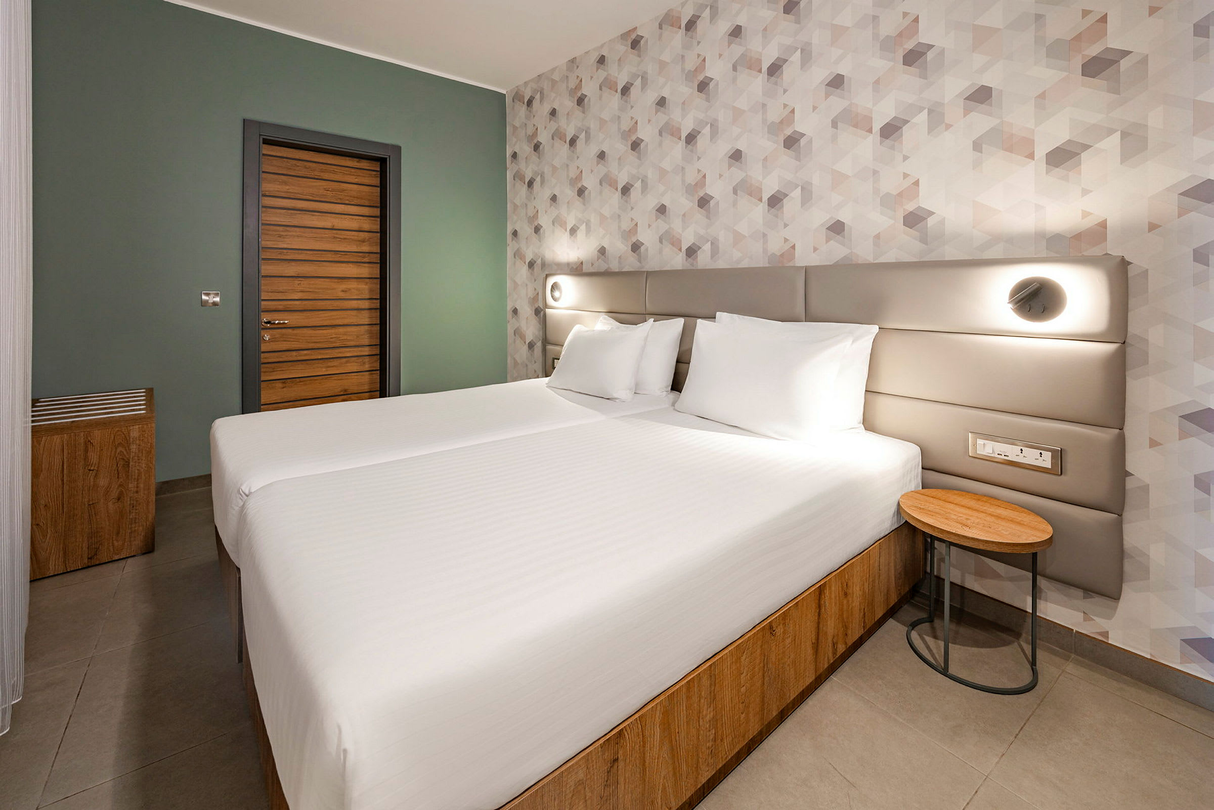 Et moderne hotellrom med to enkeltsenger som står sammen, en tredør og geometrisk mønstret tapet. (AI-generert tekst)